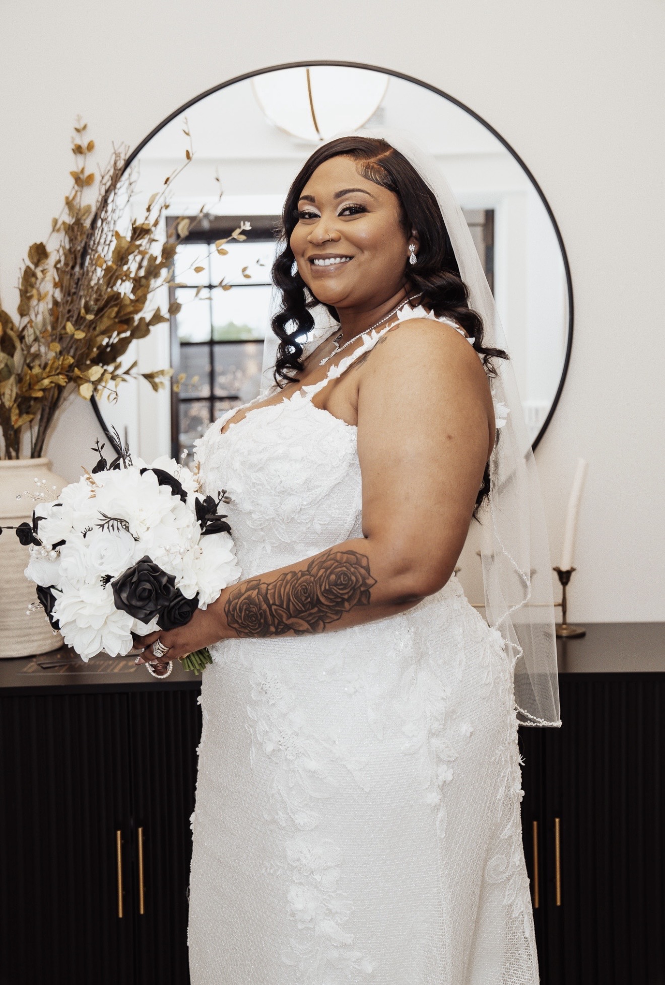 Woman in wedding dress holding bouquet, smiling, standing indoors with mirror and decorative plants behind her.