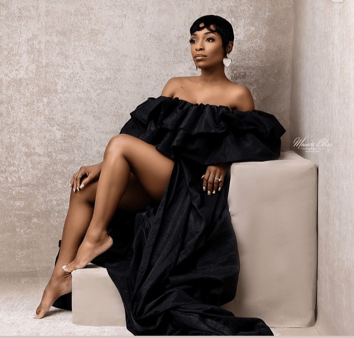 Woman with short hair in an off-shoulder black dress sitting on a white block against a textured wall.