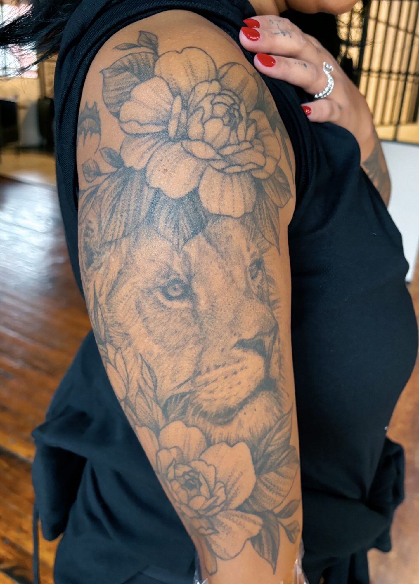 Tattoo of a lion's face surrounded by large flowers on person's upper arm, with red nail polish and a ring visible.