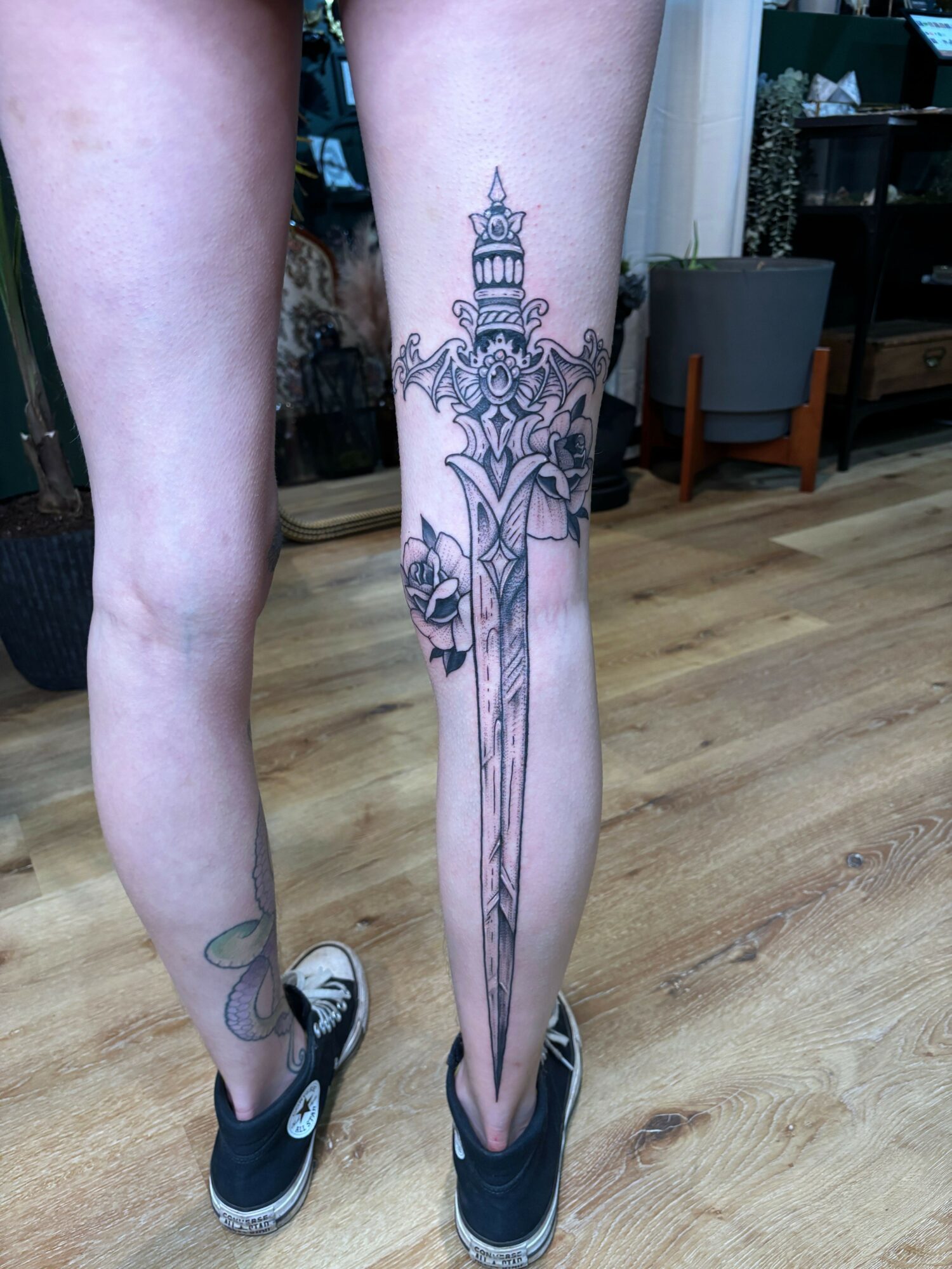 Person with a large, detailed tattoo on their leg, wearing sneakers, standing on a wooden floor.
