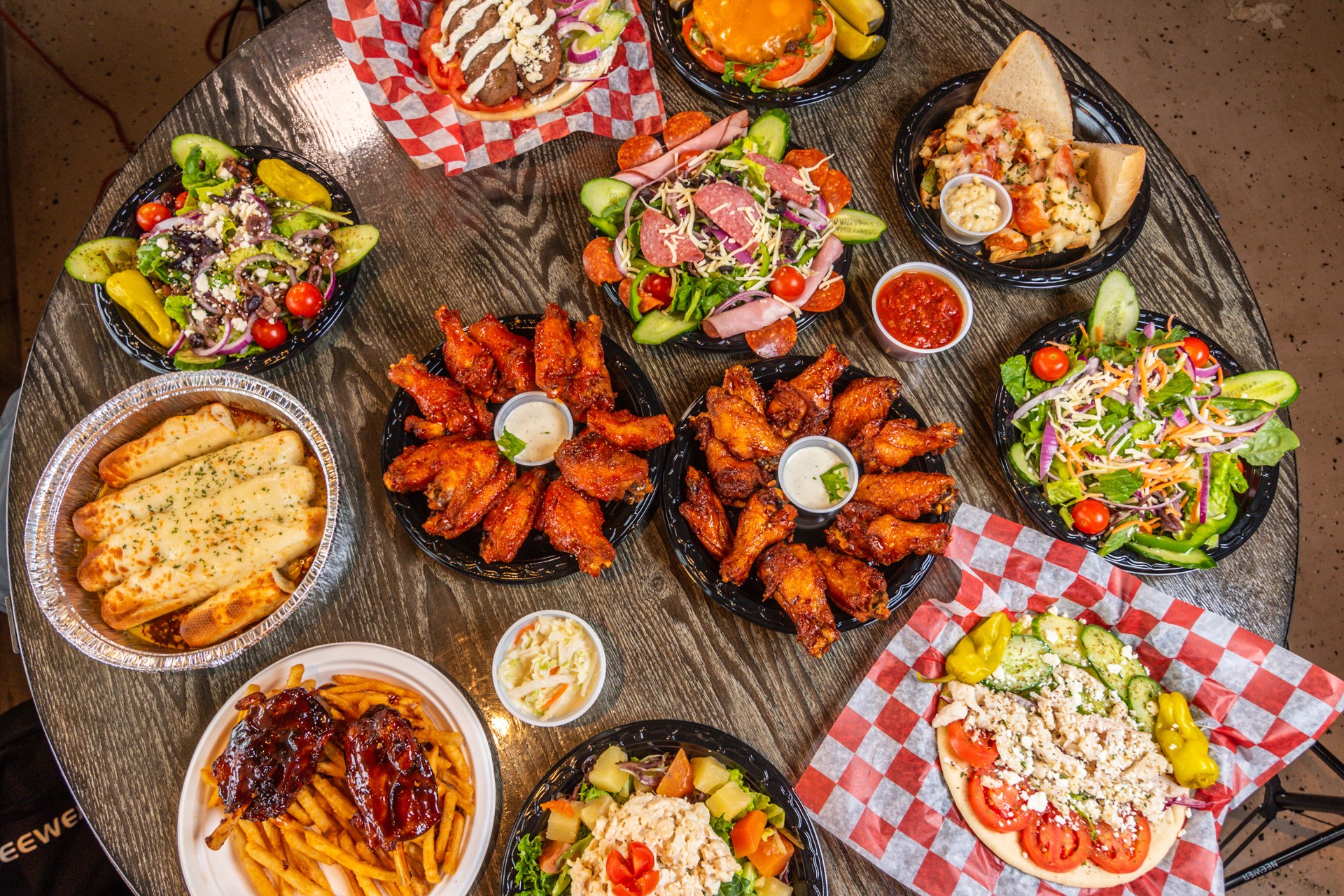 Assorted dishes including salads, fried chicken, bread, and dips arranged on a round table with red checkered paper, viewed from above.