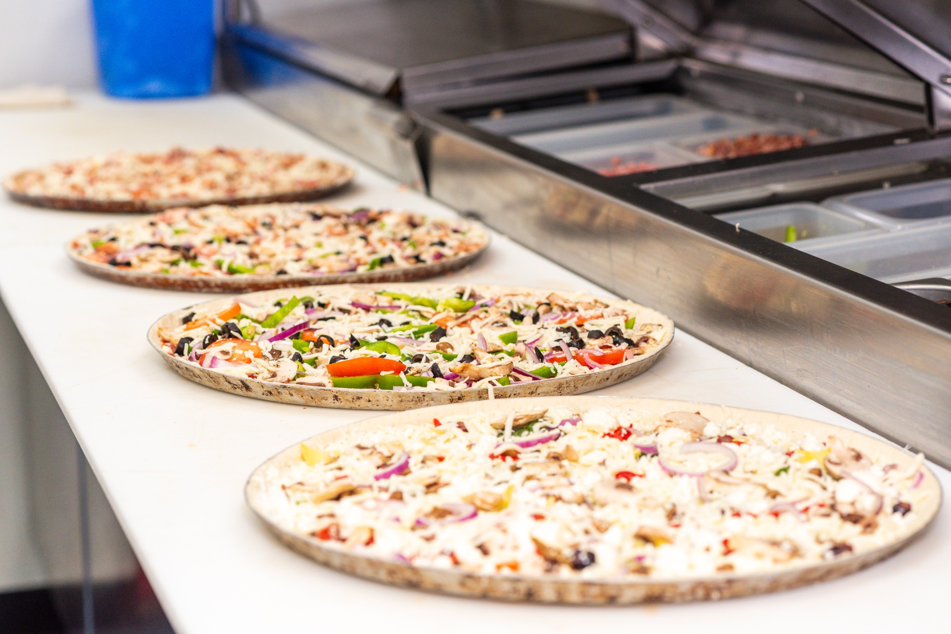 Four uncooked pizzas on a white counter, with toppings like vegetables and cheese, near a pizza oven.
