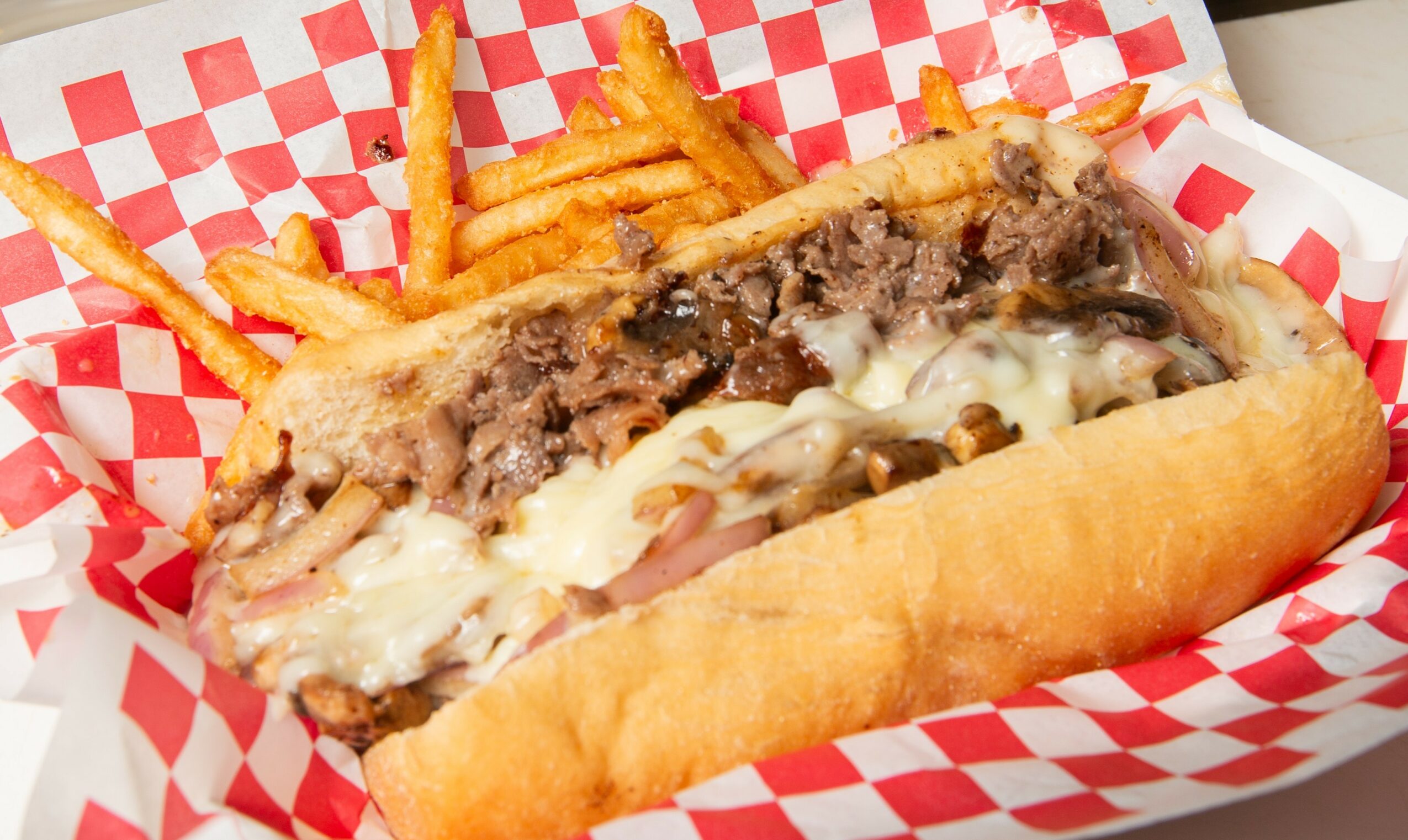 Sub sandwich with melted cheese, ground beef, and fries in a red and white checkered paper tray.