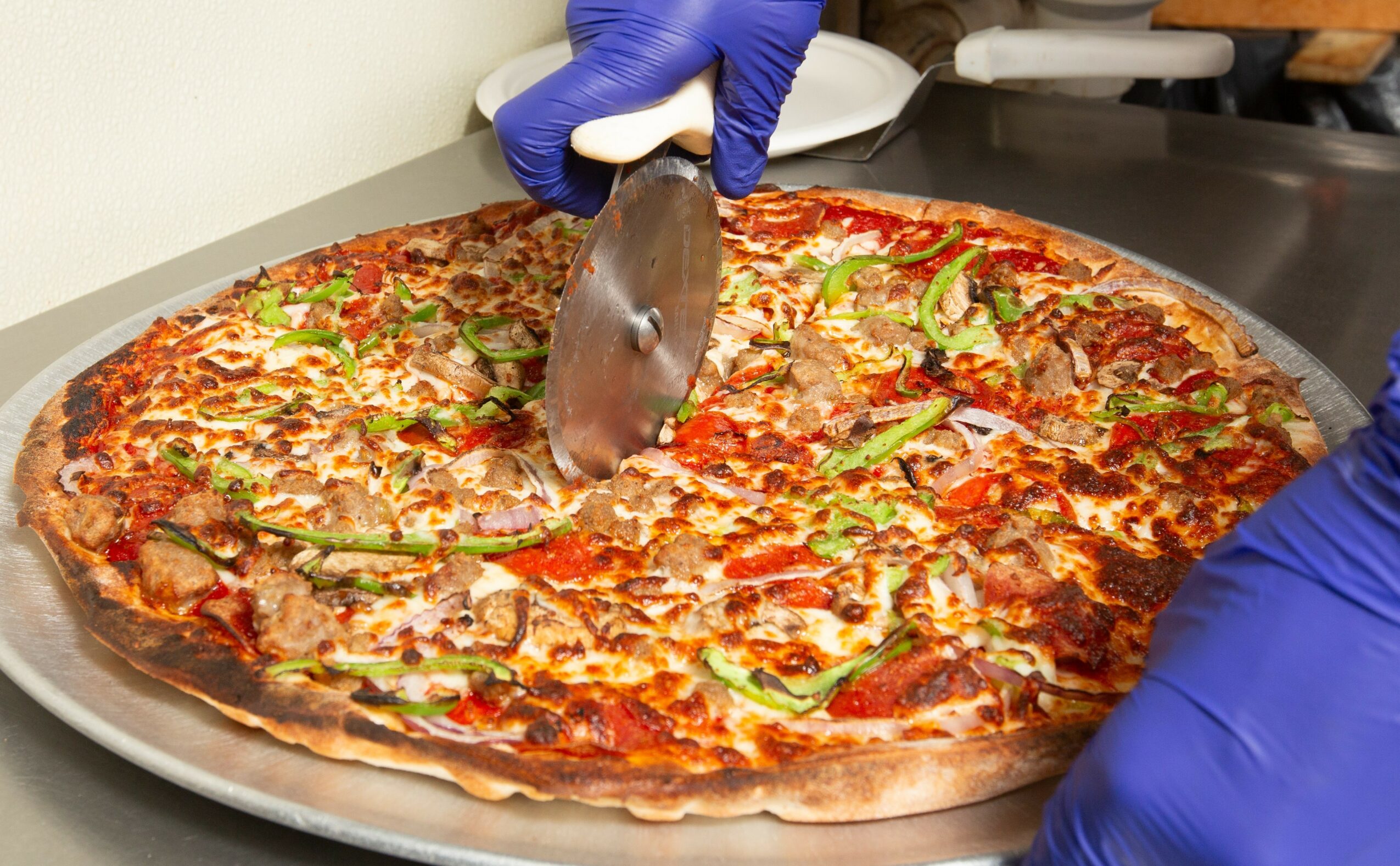 Person wearing blue gloves slicing a large pizza with toppings including green peppers and meat on a metal tray.