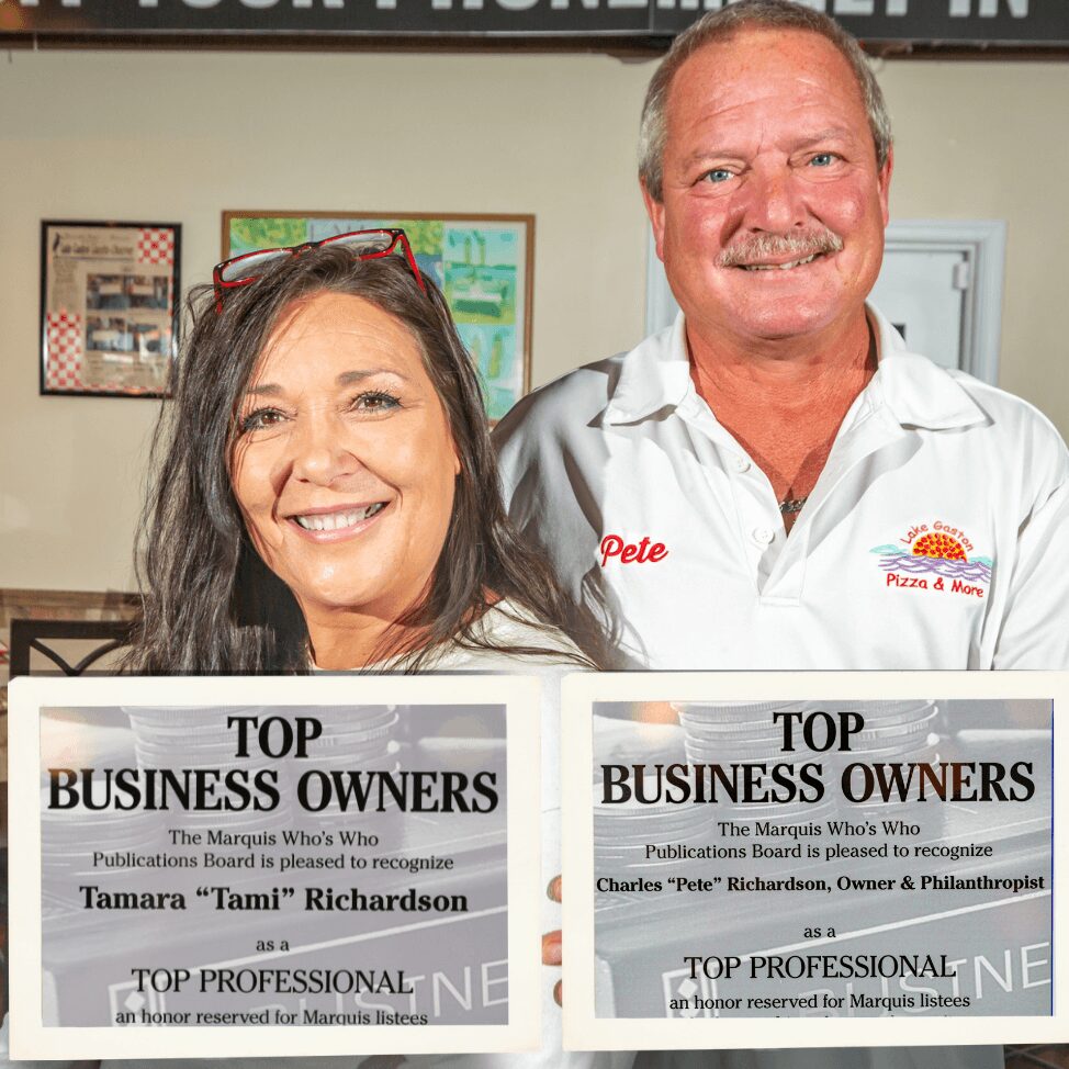 Two smiling people, a woman with dark hair and a man with a mustache, standing indoors. The woman wears glasses on her head. They hold signs recognizing business owners. The woman’s sign mentions Tamara 'Lami' Richardson. The man wears a white shirt with a logo and the name 'Pete'.