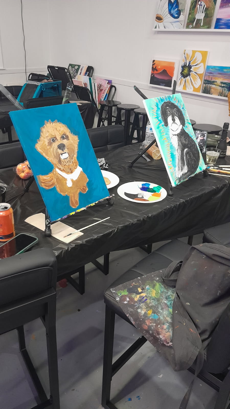 Paintings of animals and a person displayed on a table in an art studio, with chairs and art supplies visible.