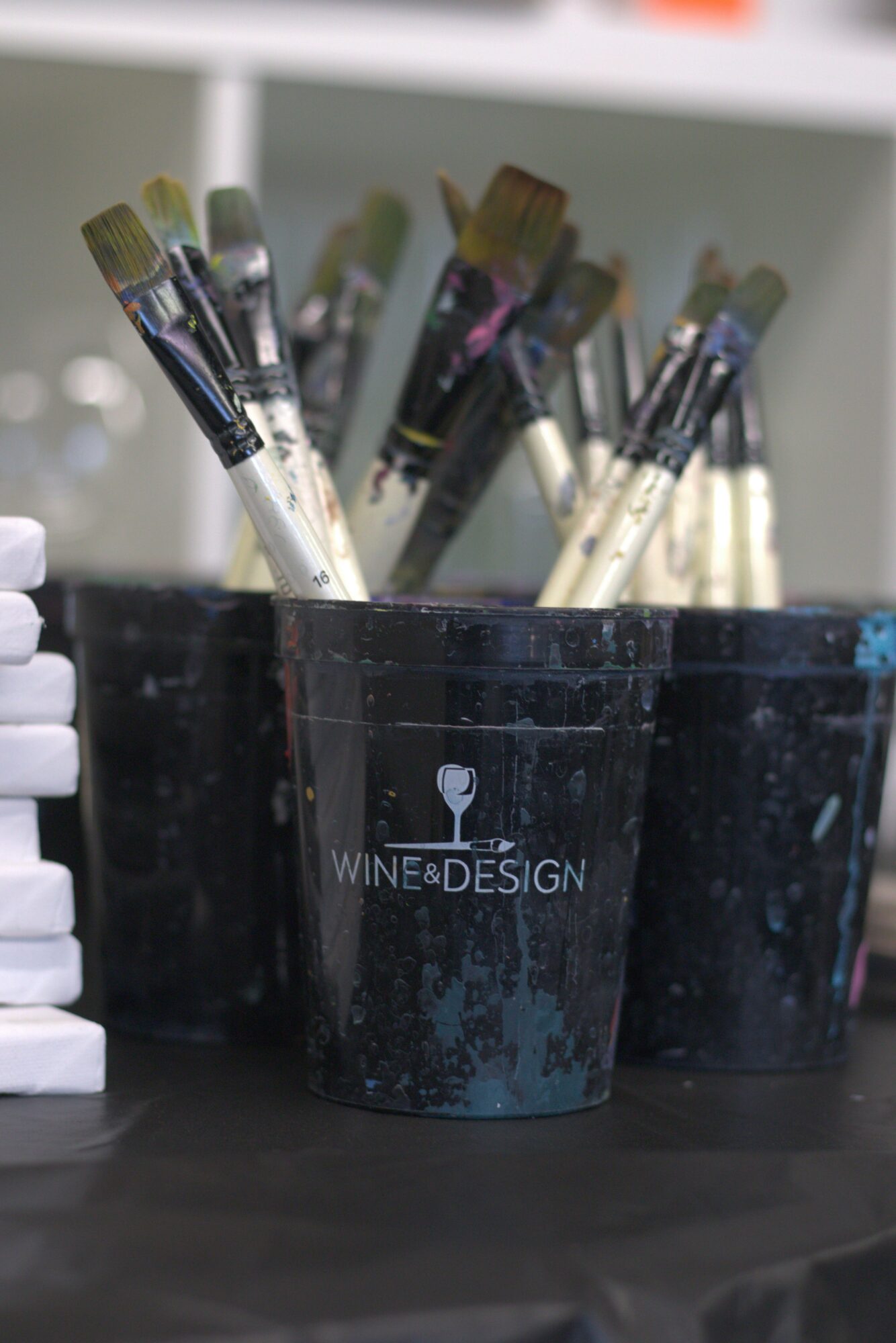 Black cup filled with paintbrushes, with a logo reading 'Wine & Design' and a wine glass icon, on a table.