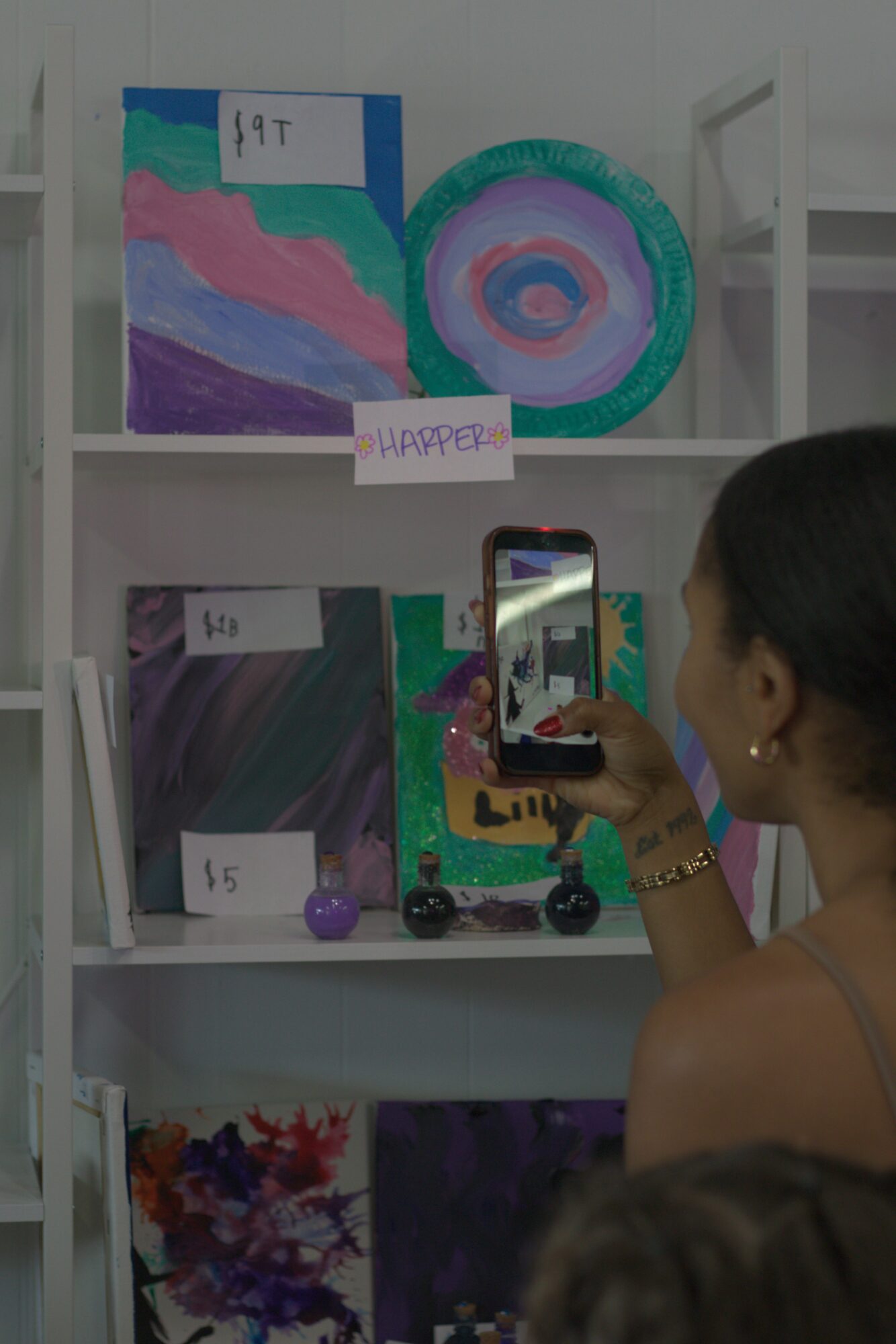 Person taking a photo of colorful artwork on a shelf, with paintings and a smartphone visible.