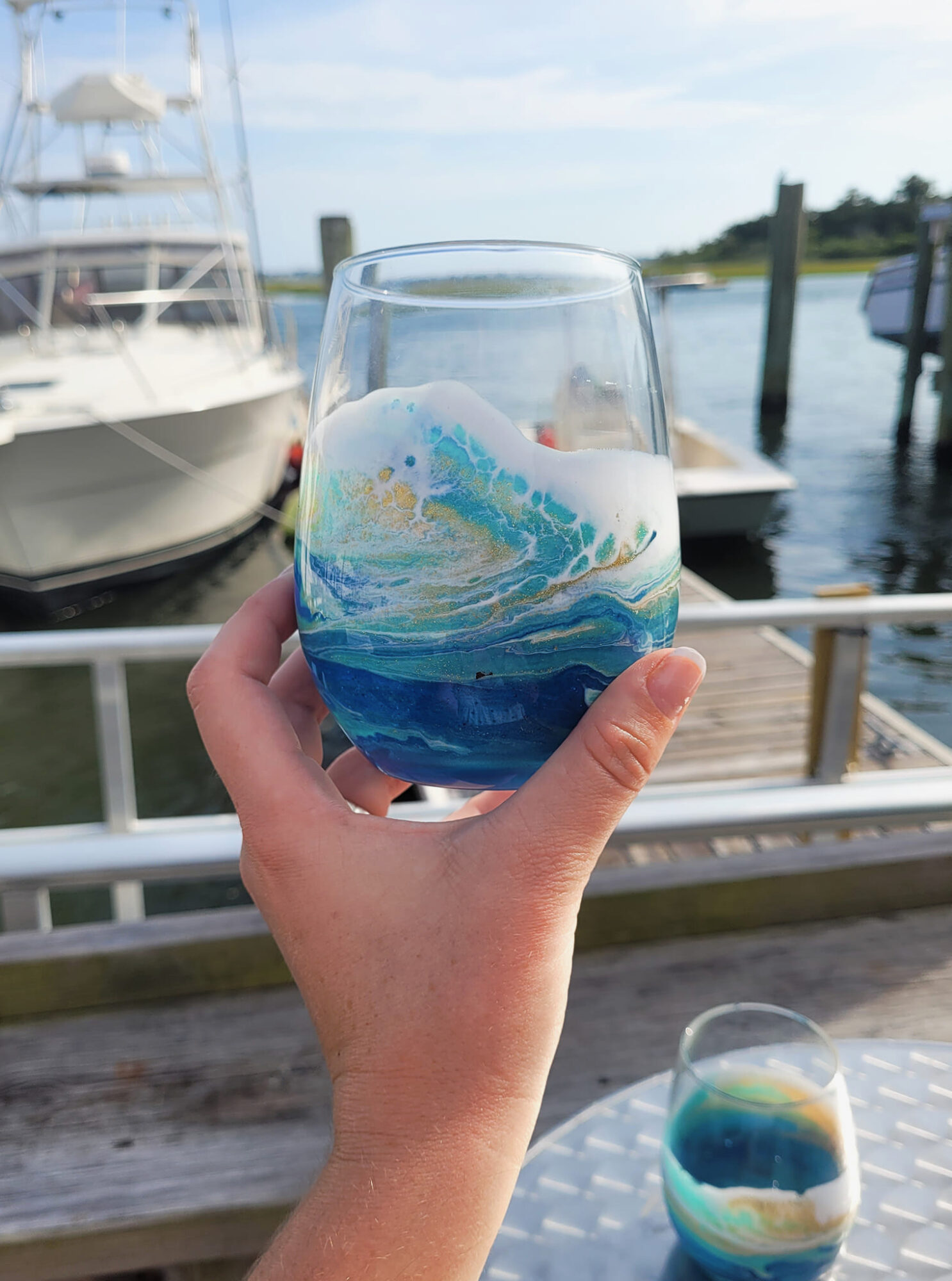 Hand holding a glass with blue and white swirling design, dock and boats in background, water and sky visible.