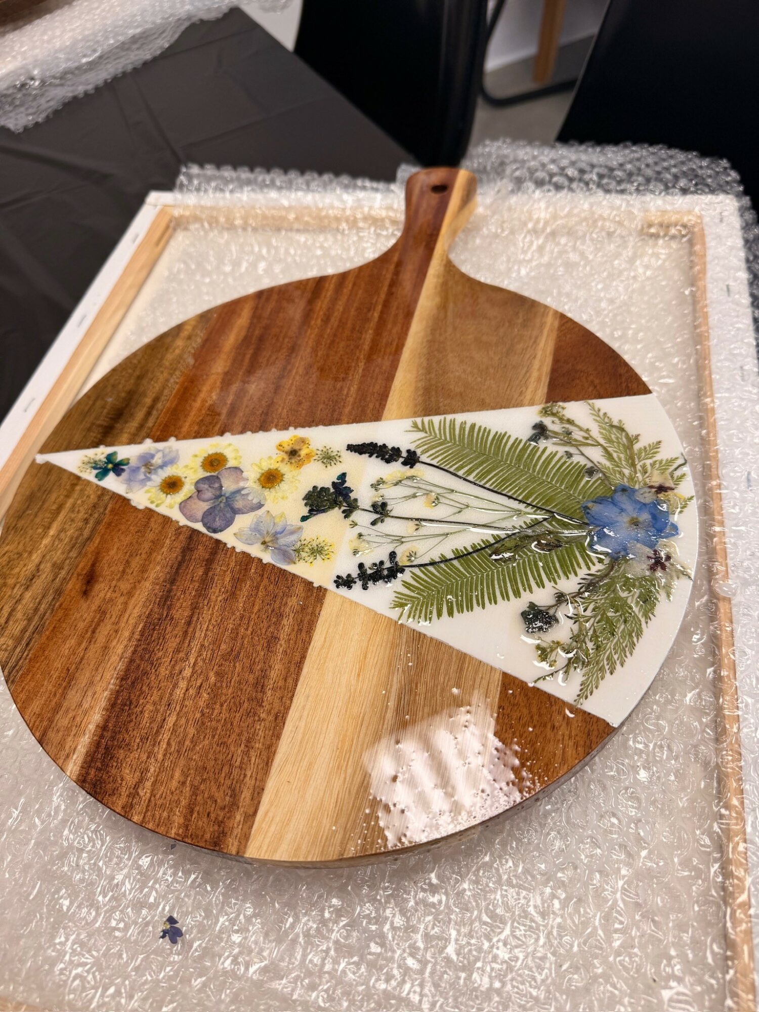 Decorative wooden paddle with a floral design on a white background, placed on bubble wrap and a black tablecloth.