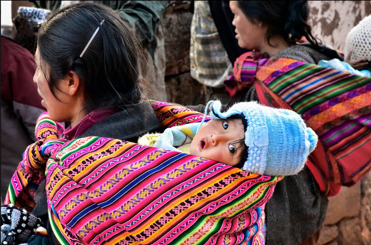 Child in a blue hat peeks out from a colorful woven cloth carried by a woman, in a crowded outdoor setting.
