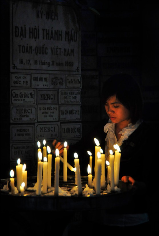 A young girl with dark hair and a white shirt lights candles in a dark setting, with a poster in the background.