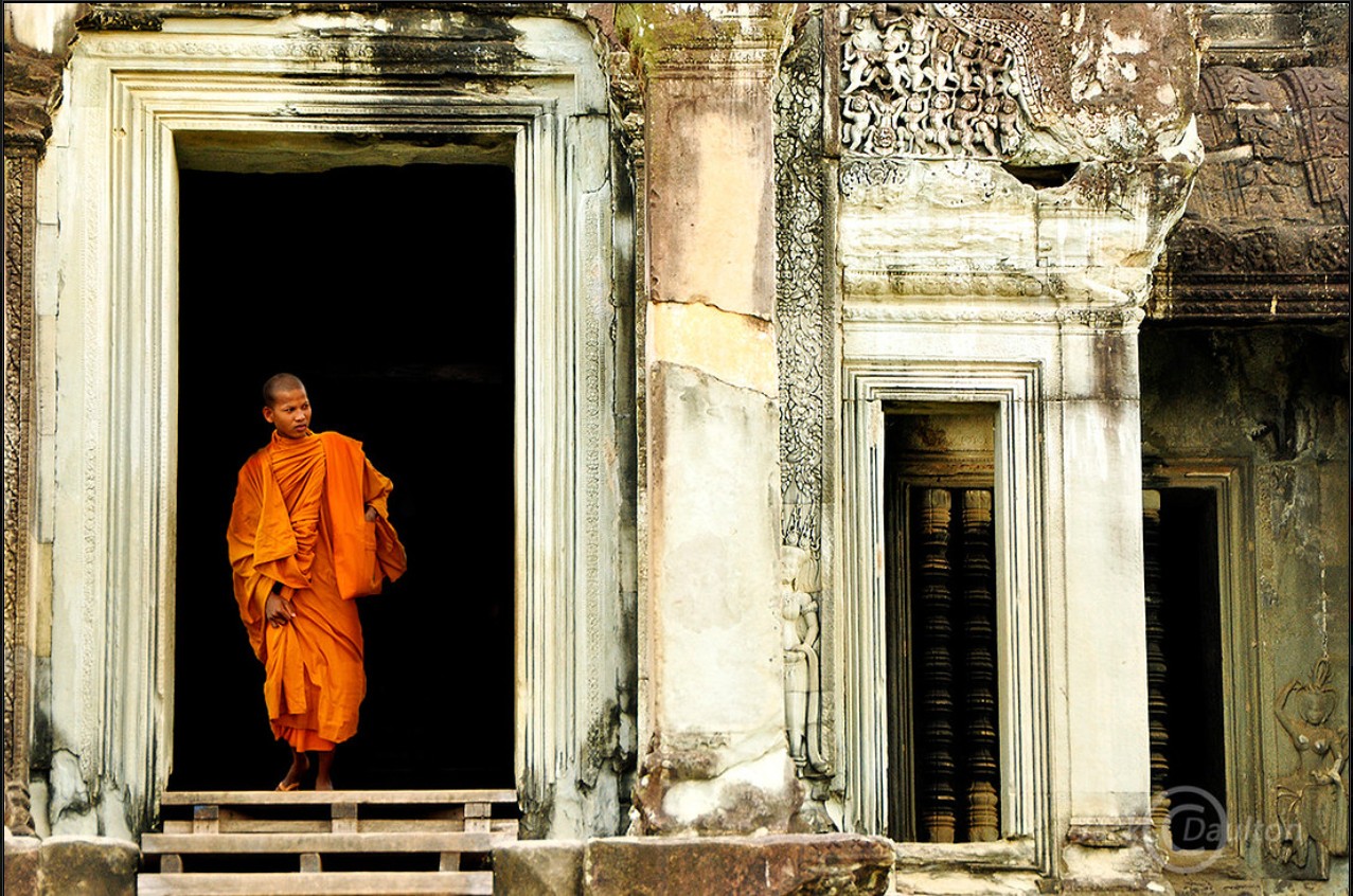 A monk in orange robes standing in an ancient stone doorway with weathered walls.