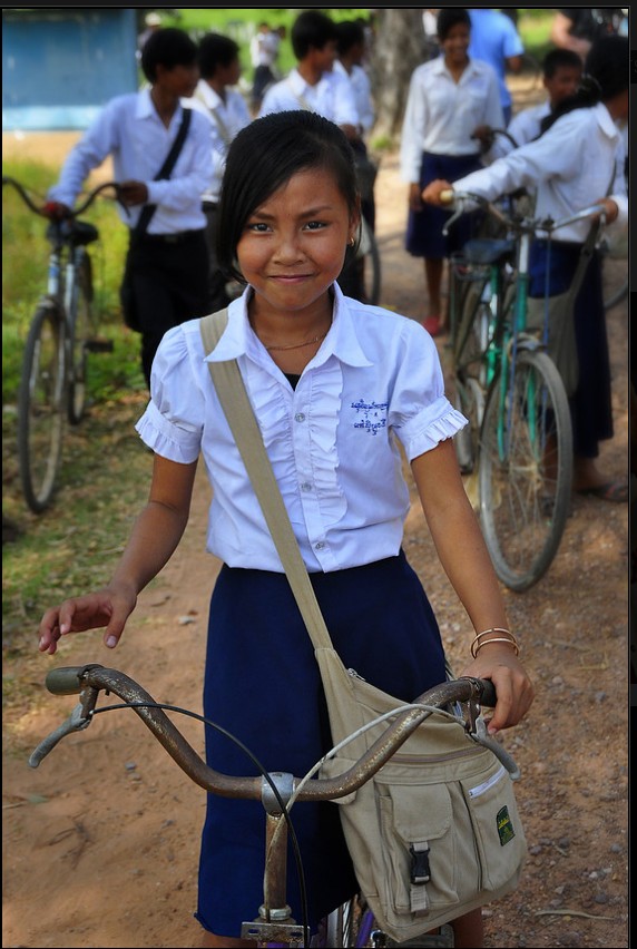 Young girl in school uniform standing outdoors with bicycles and other students in the background.