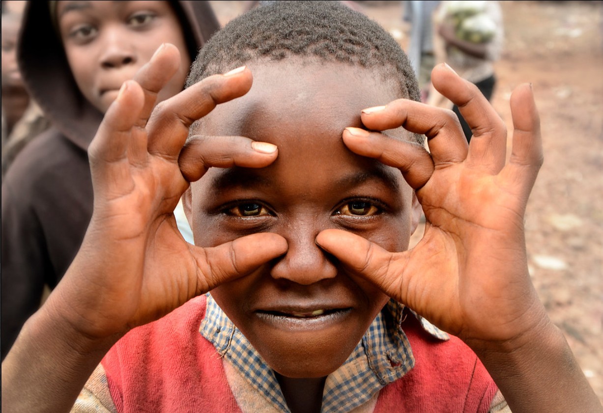 Child holding hands around face, smiling, with another child in background outdoors.