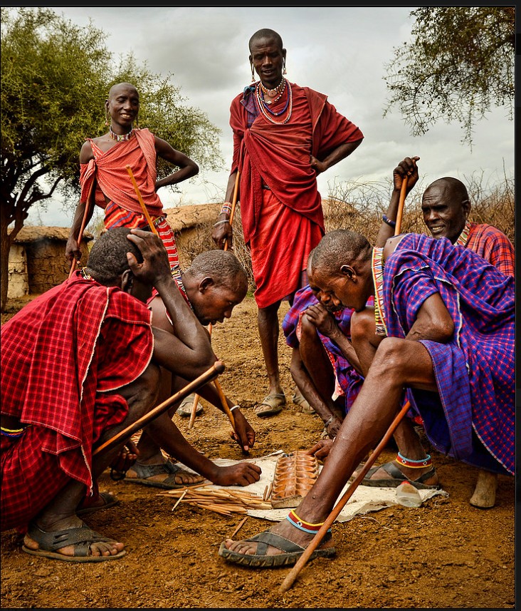 Group of people outdoors, some sitting and some standing, engaging with objects on the ground, trees in background.