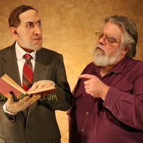 Two men engaged in conversation, one holding a book and the other pointing at him, against a plain background.