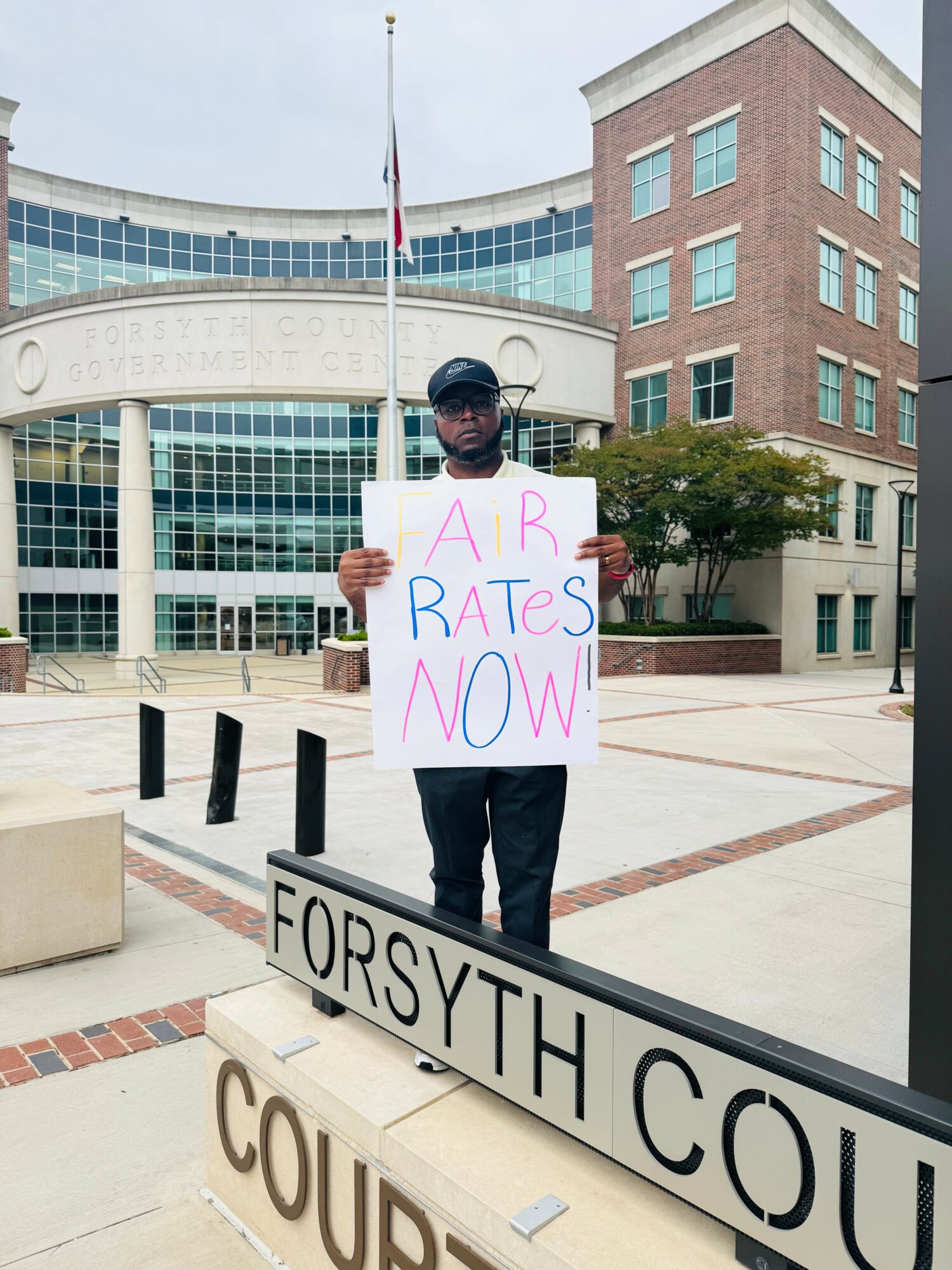 Person holding a sign that says 'AP BATES NOW!' standing outside Forsyth County building with modern architecture.