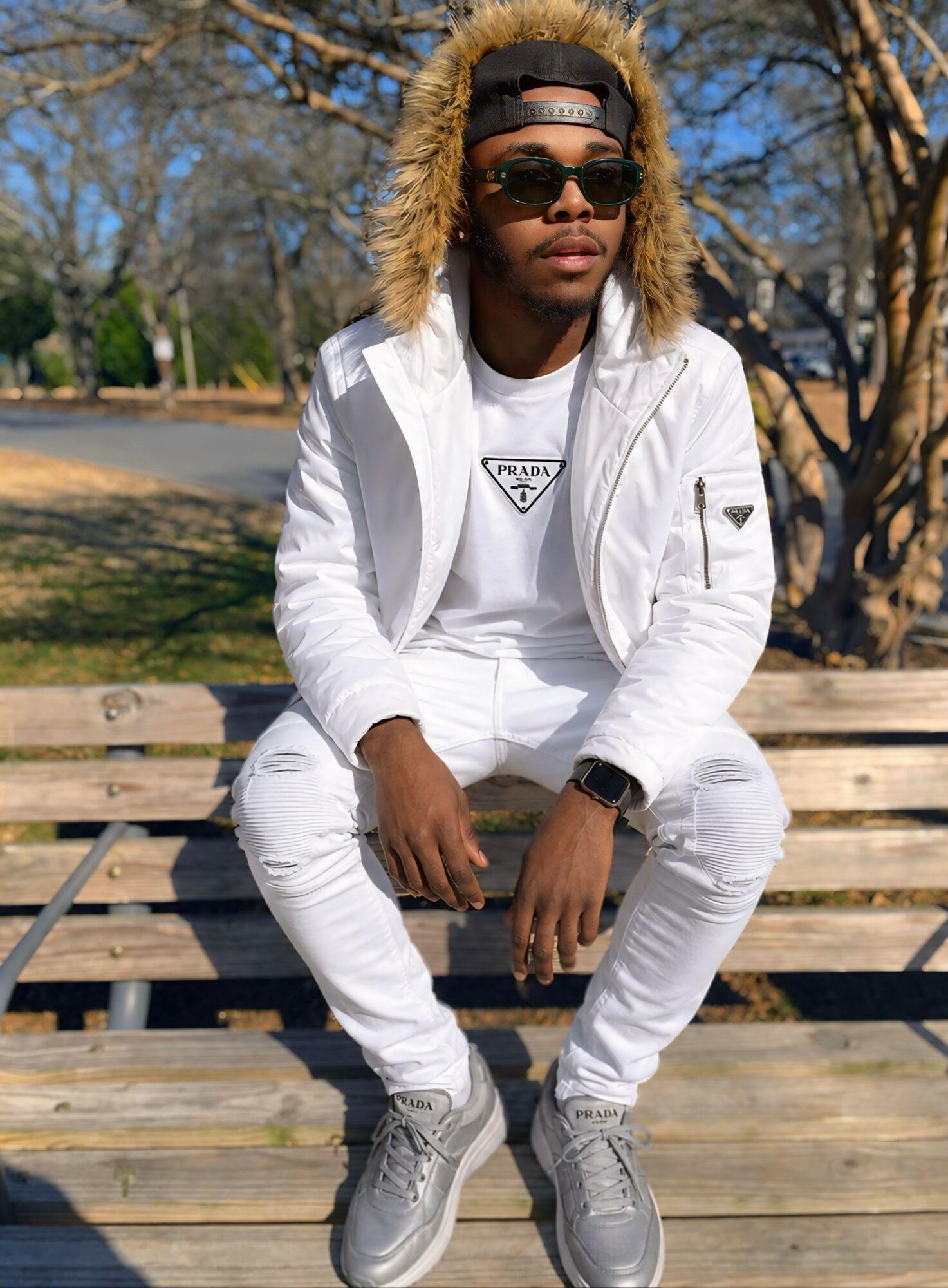 Person sitting on a park bench wearing sunglasses, a white jacket, and gray sneakers, with trees and blue sky in background.