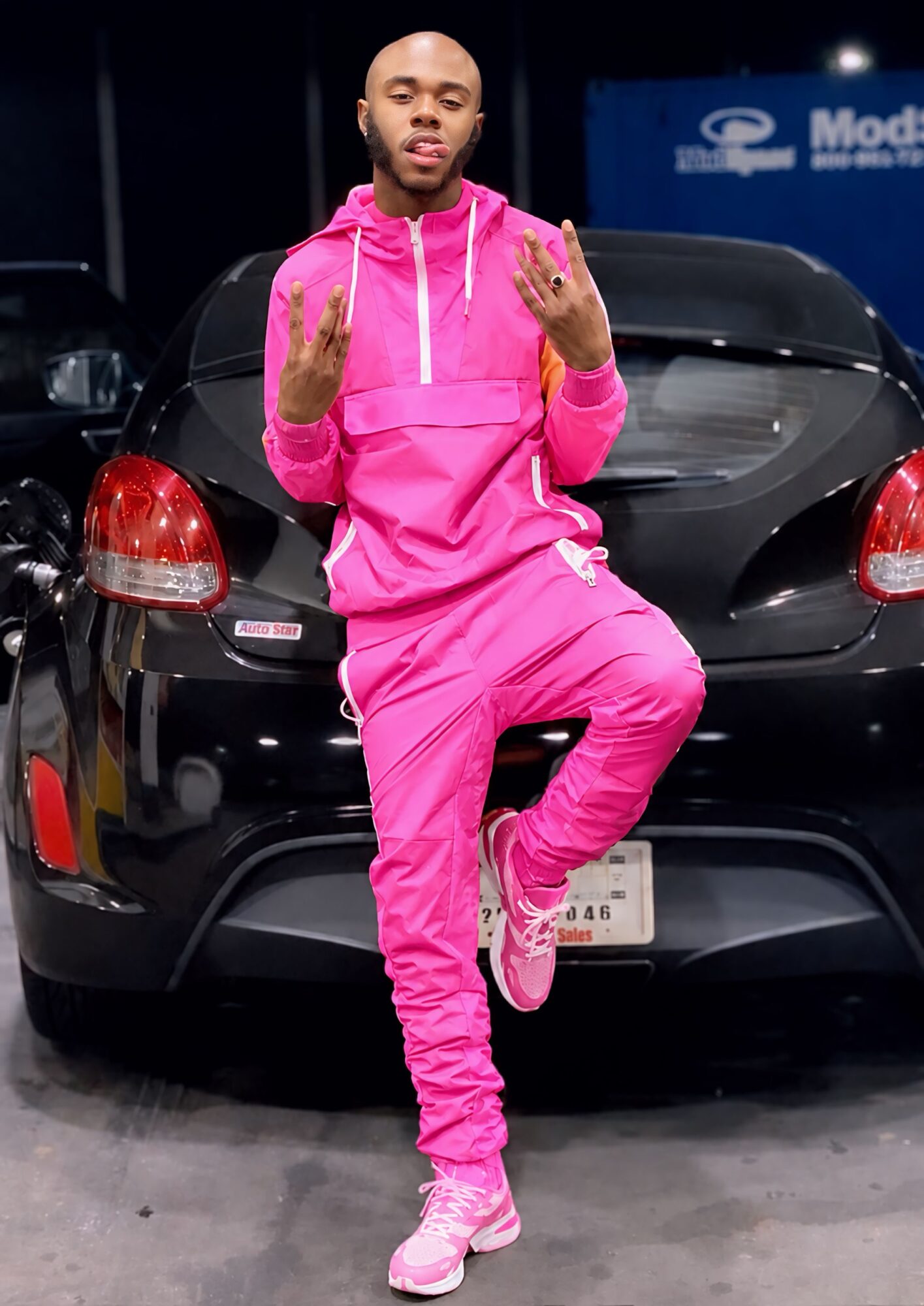 Young man in pink tracksuit standing in front of a black car, making hand gestures, indoors with a blue sign in the background.