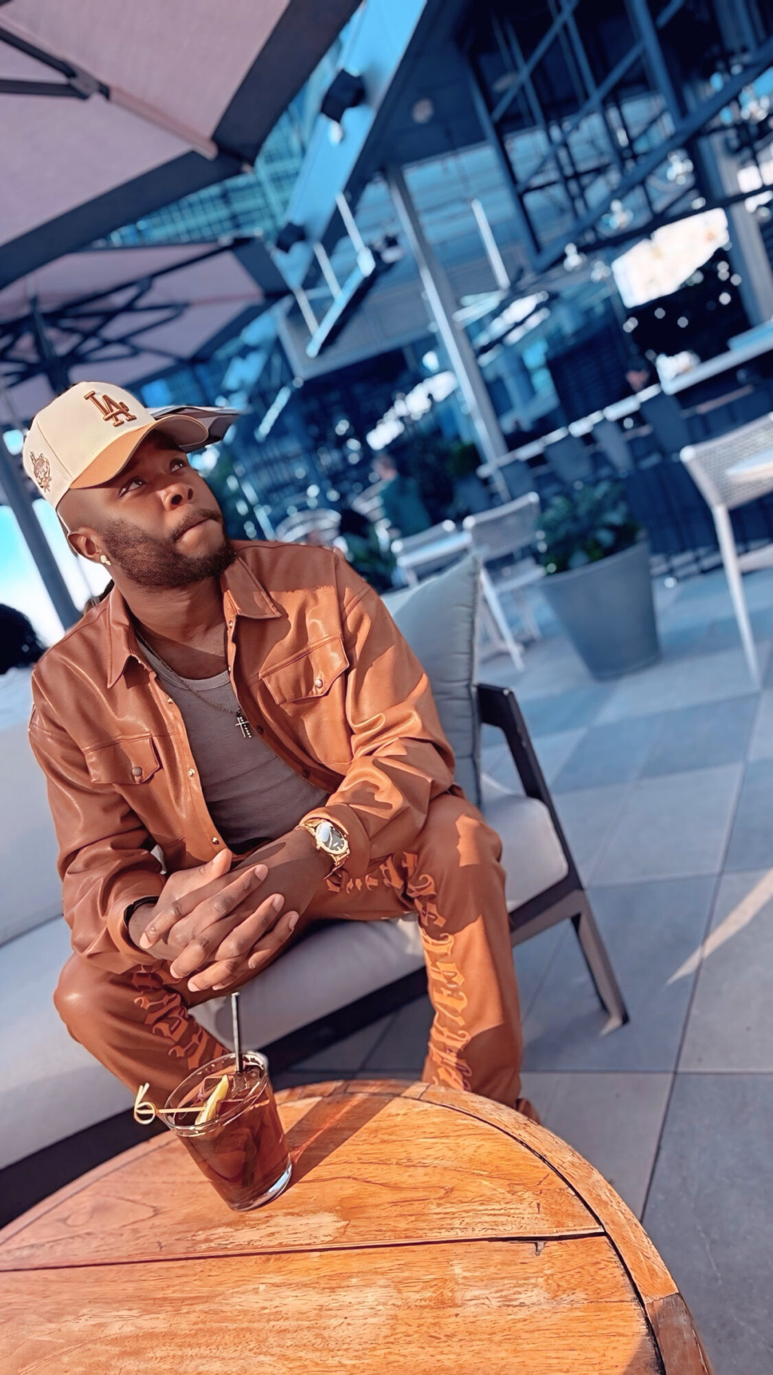 Man sitting at a table with a drink, wearing a cap and casual clothing, in an indoor setting with large windows.