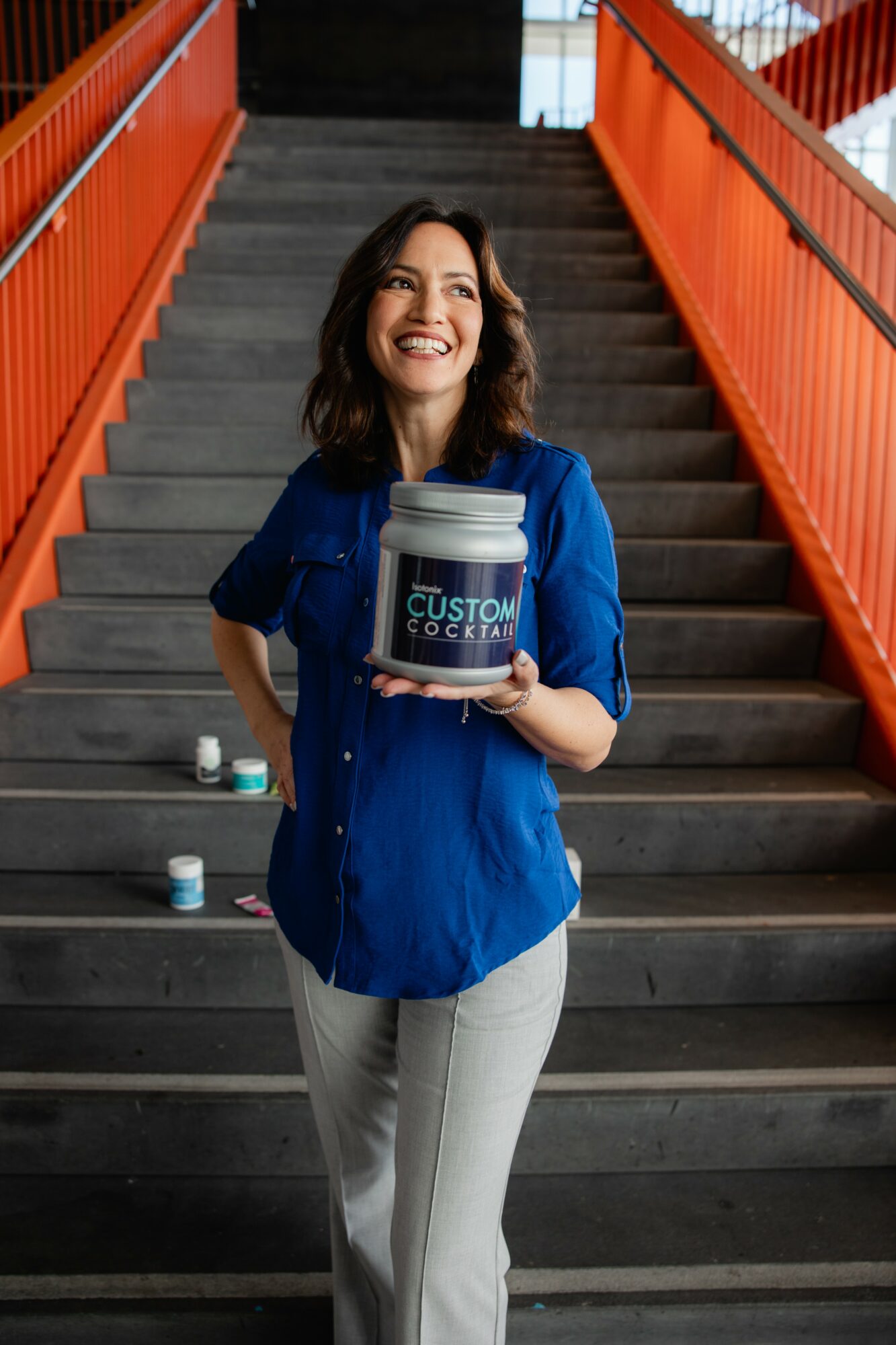 Woman smiling on stairs holding a container labeled 'Custom Cookie Co.' with orange railings in background.