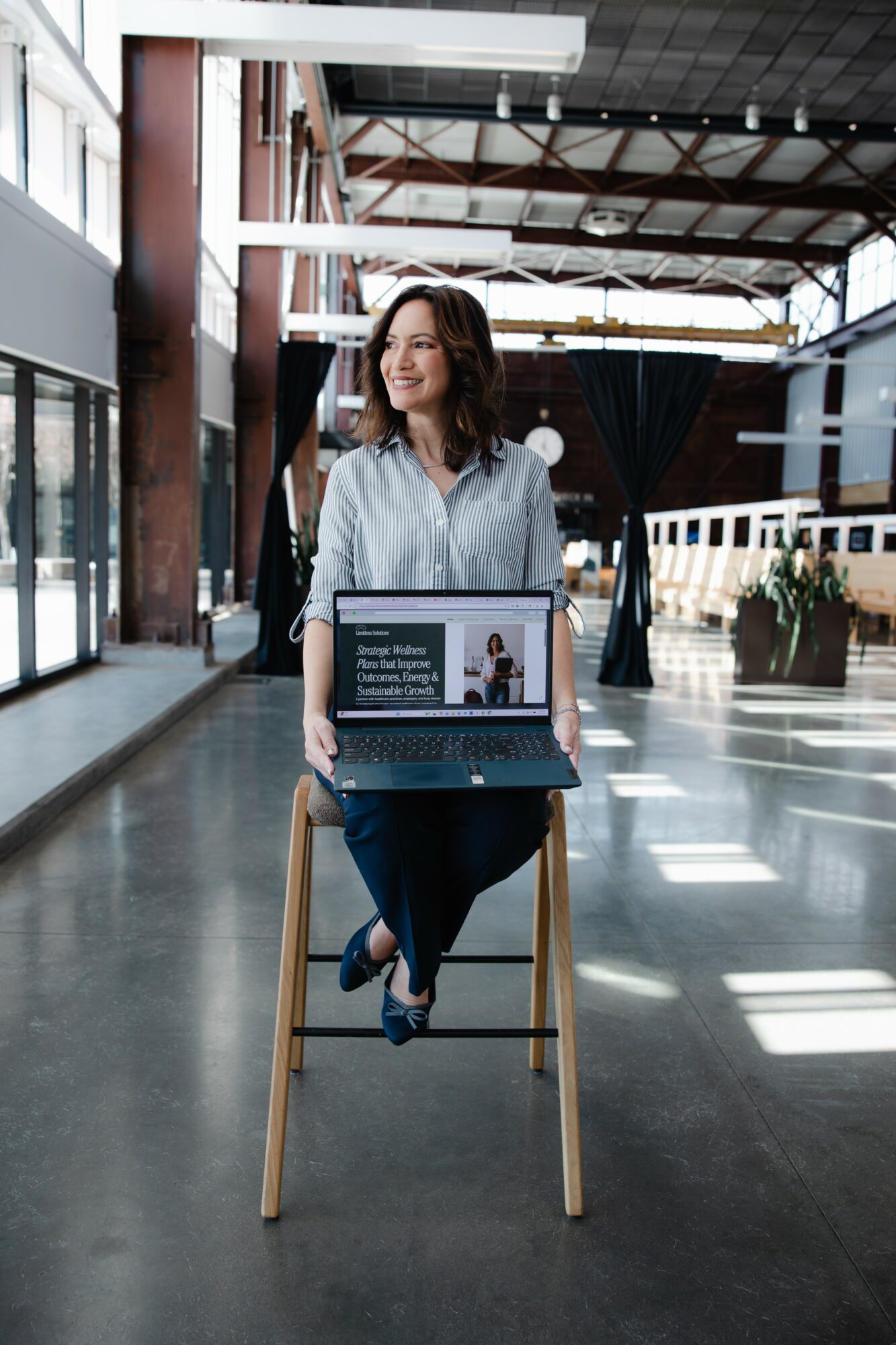 Woman sitting on a stool in a modern indoor space with a laptop on her lap, smiling and looking to the side.
