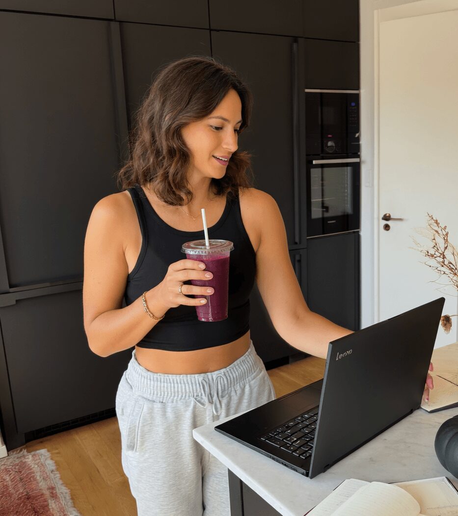Woman with shoulder-length hair in a black crop top and gray sweatpants using a laptop in a modern kitchen, holding a purple smoothie.