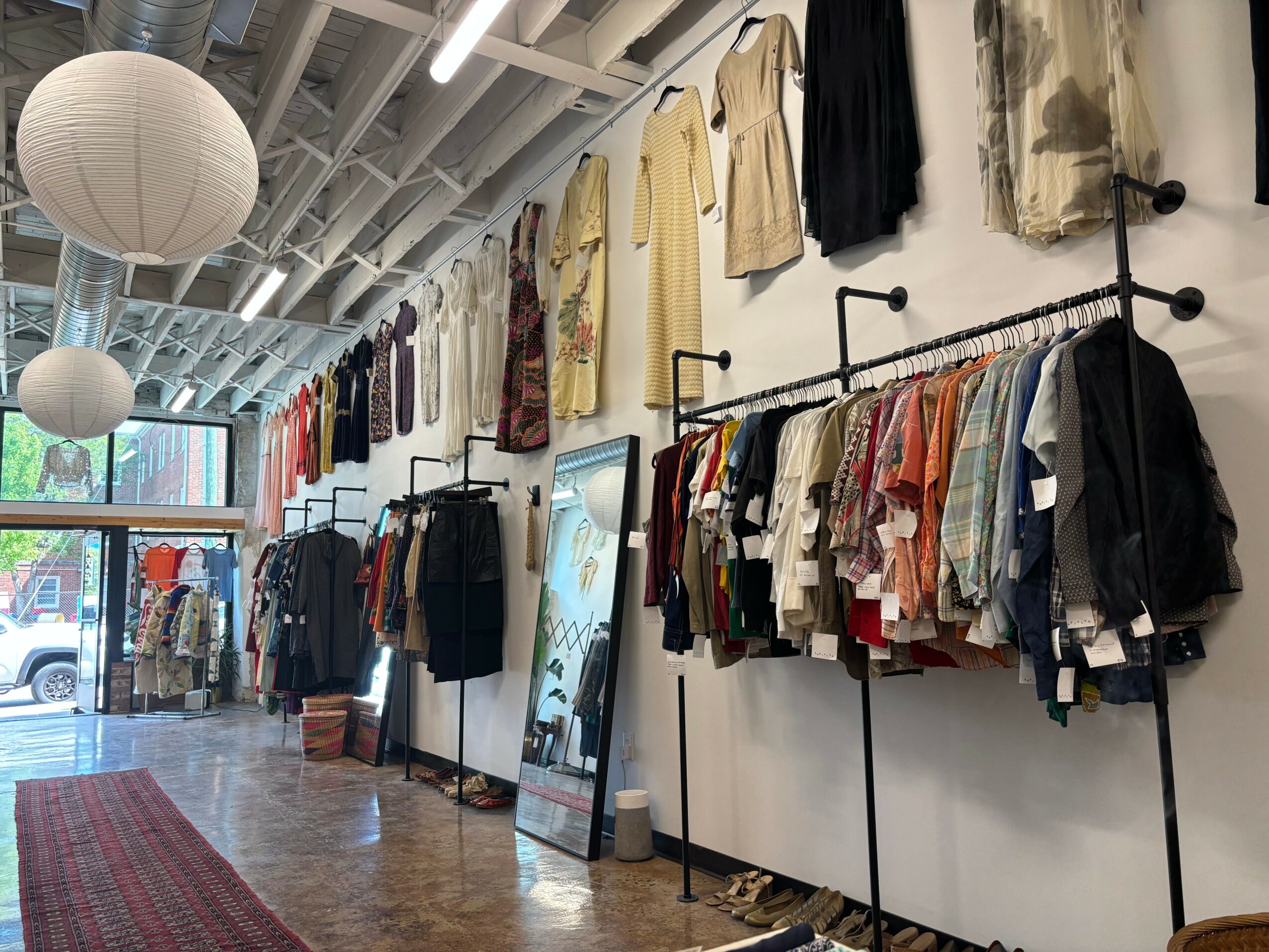 Clothing store interior with hanging garments, racks, and large windows, ceiling lights, and a red rug on wooden floor.