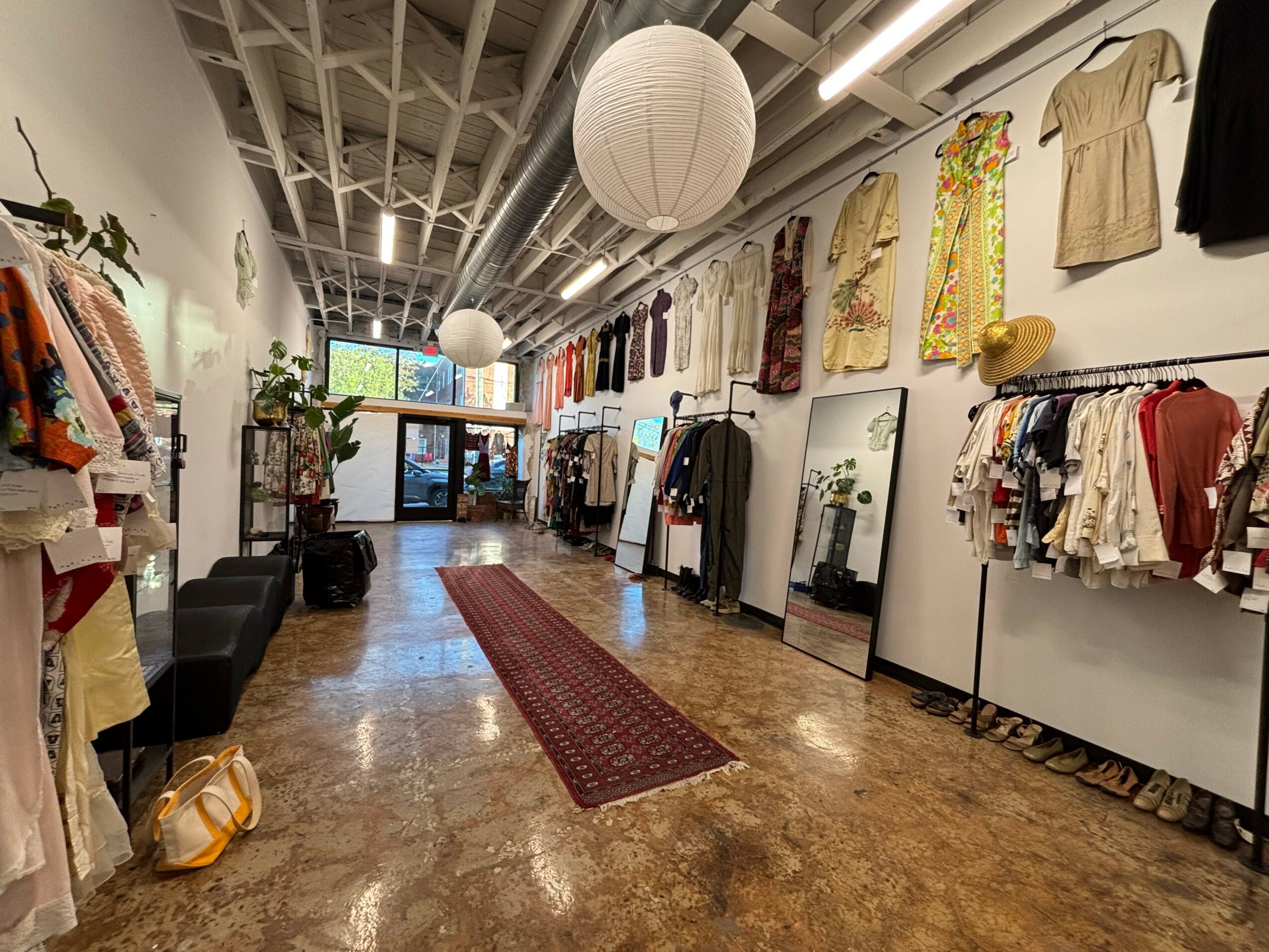 Clothing store interior with racks of clothes, mirrors, and a red rug on a wooden floor, ceiling with exposed pipes, and a paper lantern.