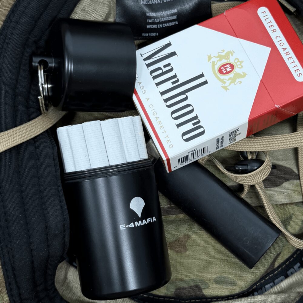 Cigarette pack, black container with white logo, and black cylindrical object on camouflage fabric.