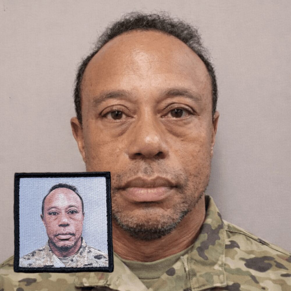 Man in camouflage shirt with a screen showing his face, neutral expression, plain background.