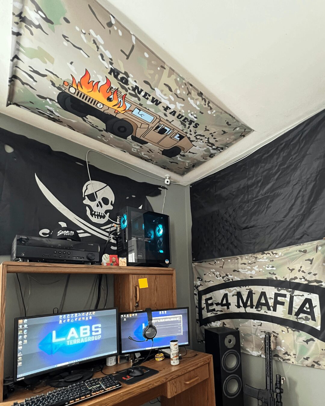 Room with a ceiling-mounted screen, a black wall with a skull and crossbones flag, and a wooden desk with two monitors, speakers, and a computer.