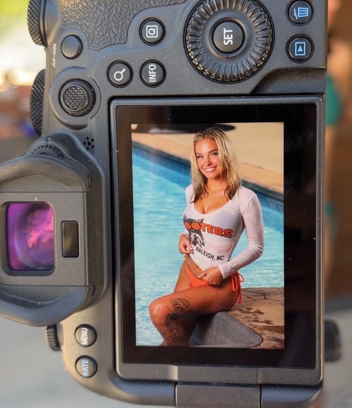 A woman sitting by a pool, smiling, wearing a white long-sleeve shirt and red shorts, with water in the background.