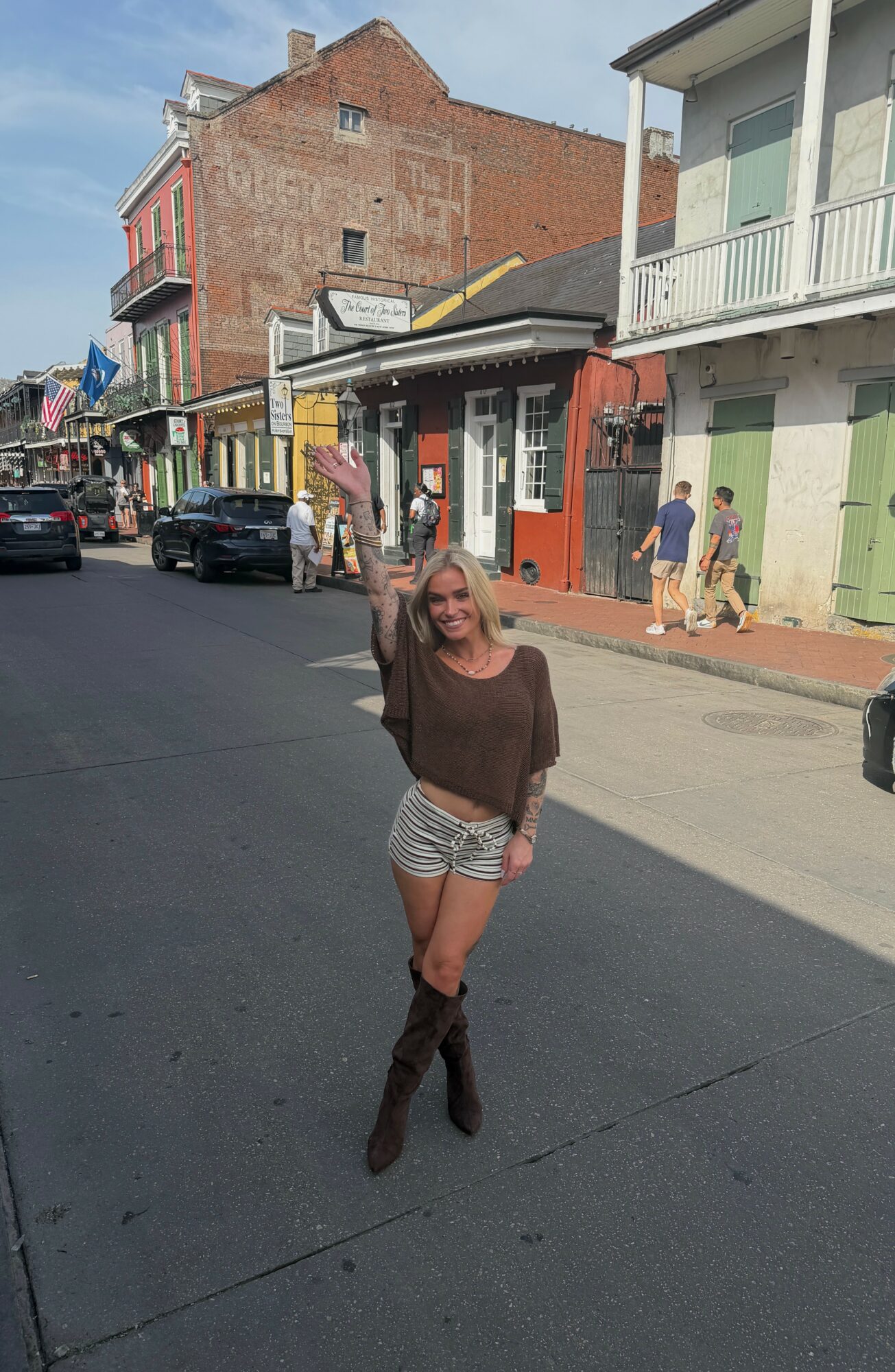Woman standing on street with buildings and people in background, smiling, waving, wearing brown top and striped shorts.