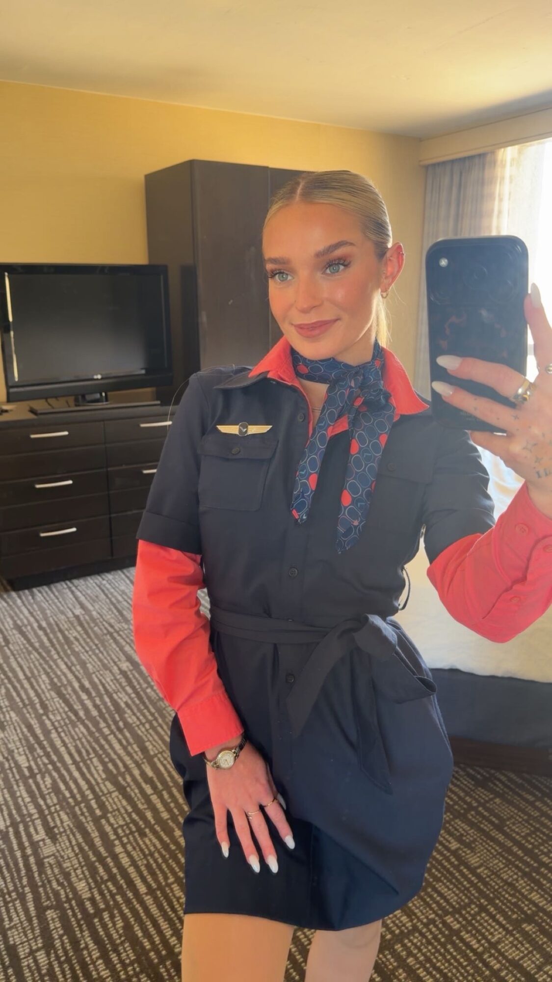 Woman in uniform taking a mirror selfie in a hotel room, smiling, with a TV and window in the background.
