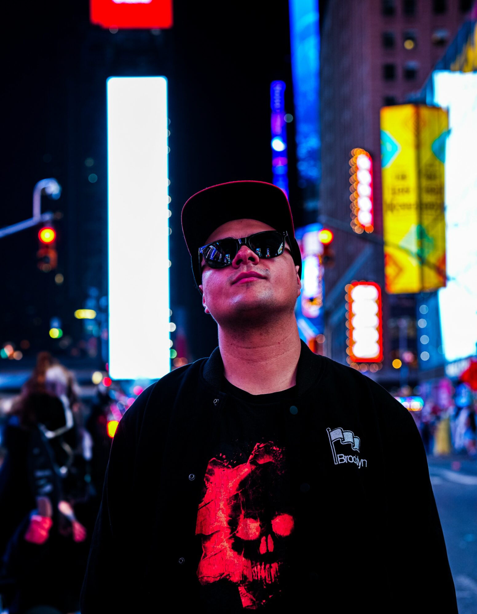 Young man wearing sunglasses and a cap stands in a brightly lit city at night with colorful neon signs.