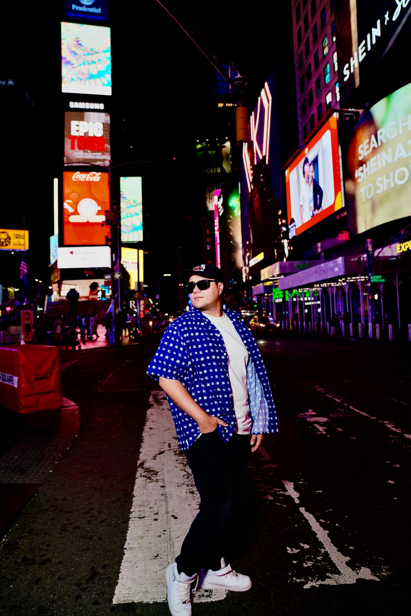Person wearing sunglasses, blue polka dot shirt, and white sneakers standing in a brightly lit city street at night with colorful billboards.