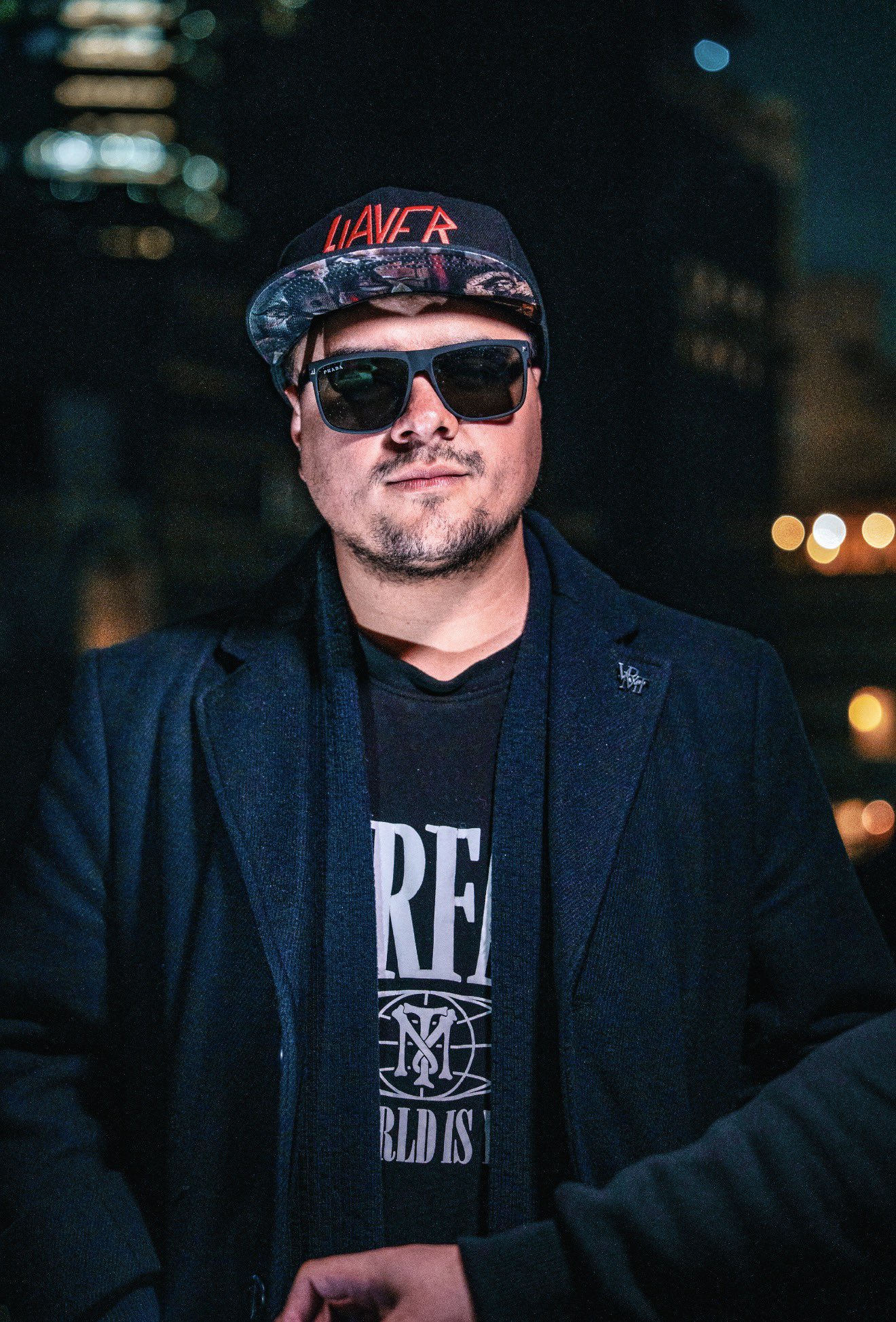 Man wearing sunglasses, cap, and dark jacket outdoors at night with city lights in background.
