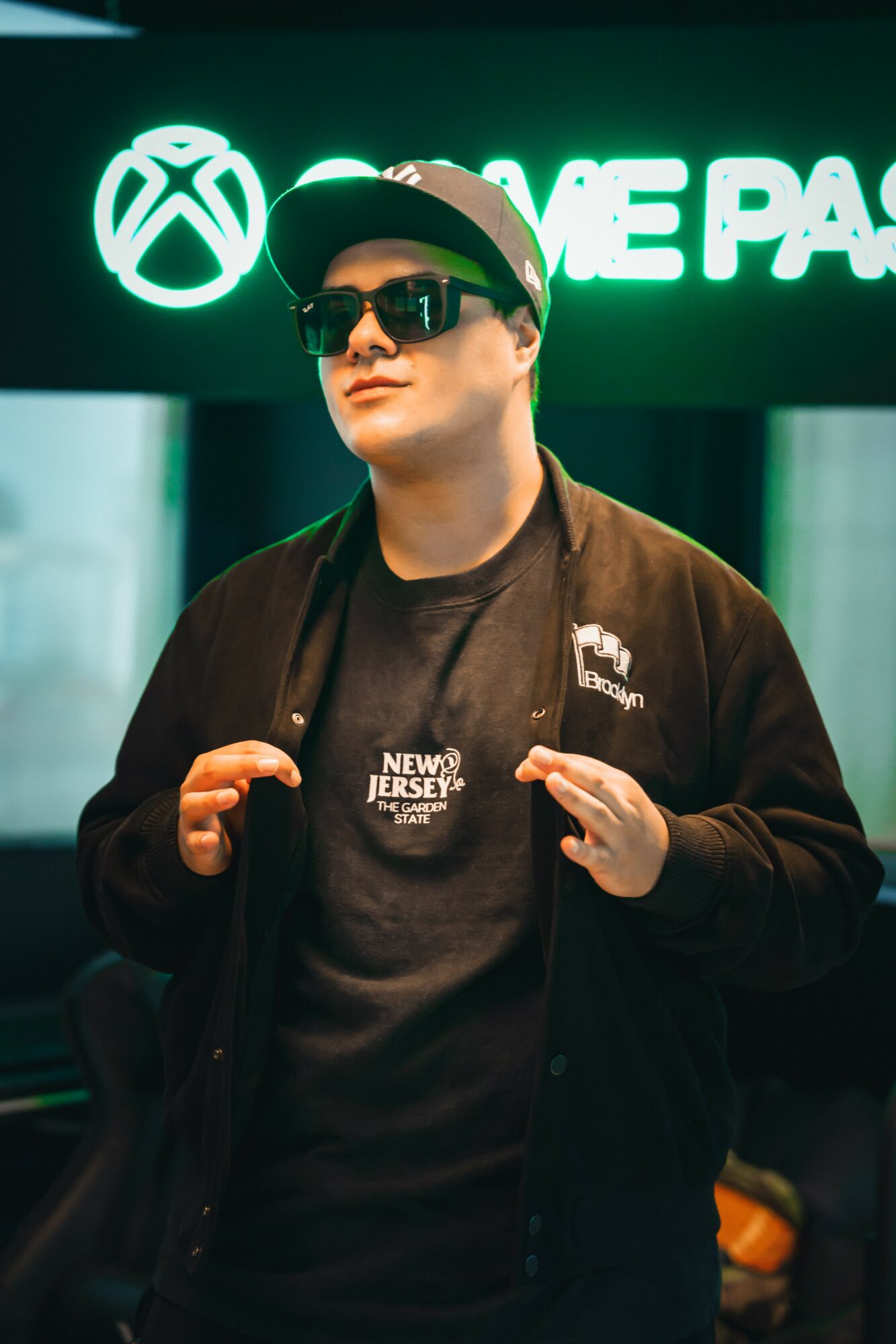 Young man wearing sunglasses, a cap, and a jacket, standing in front of an Xbox sign at a gaming event.