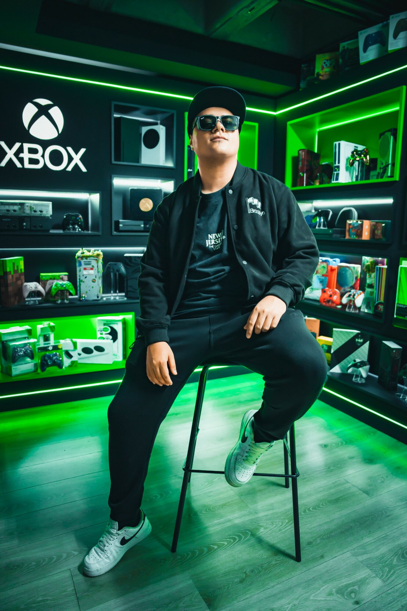 Person sitting on a stool in a gaming store with Xbox and gaming accessories, illuminated by green lighting, wearing sunglasses and a black jacket.