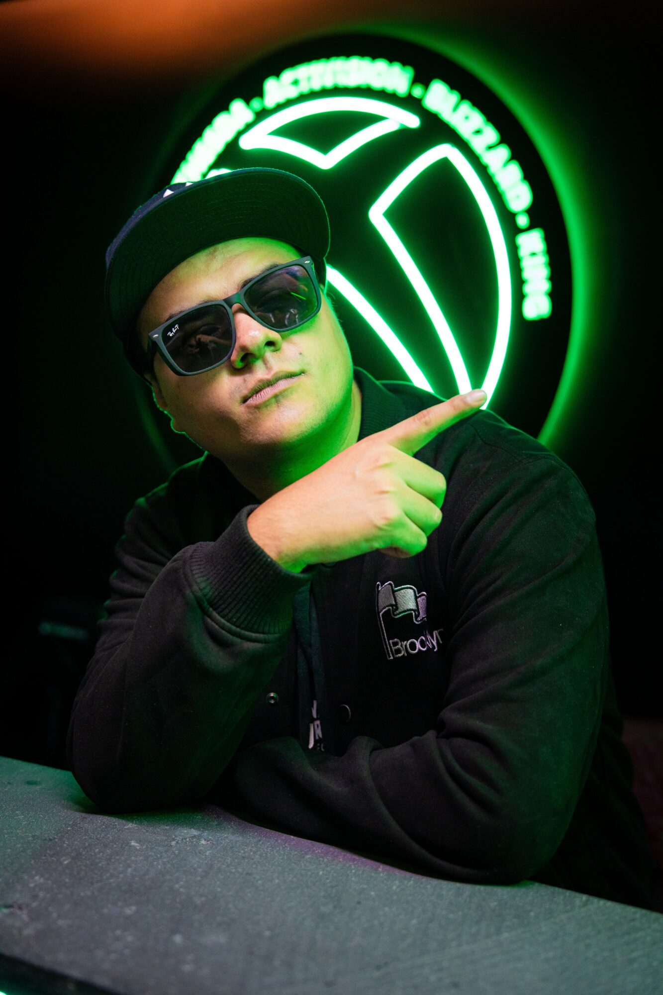 Person wearing sunglasses and a cap points at a neon green circular logo behind them.