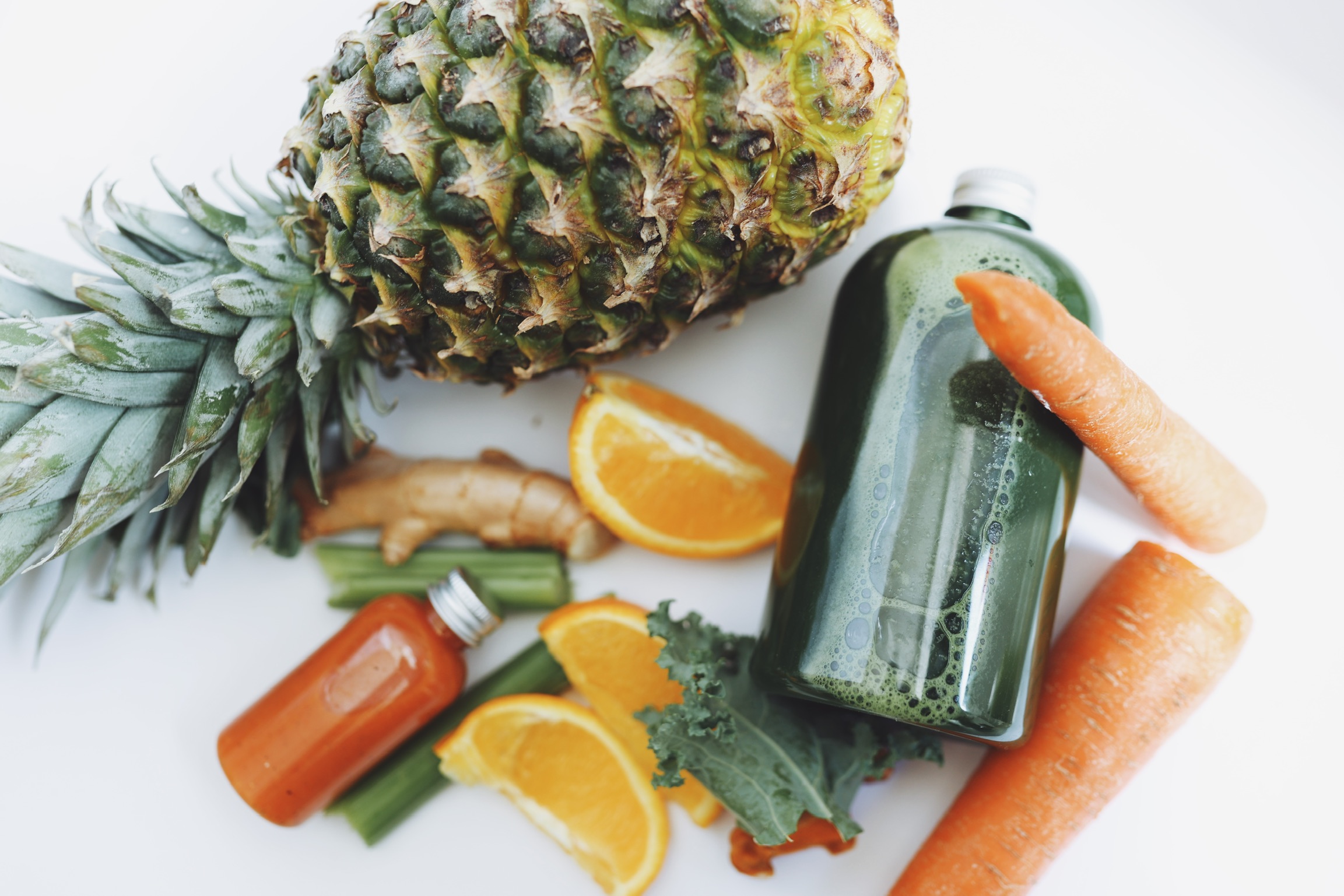 Pineapple, orange slices, a green juice bottle, carrots, ginger, and kale on a white surface.