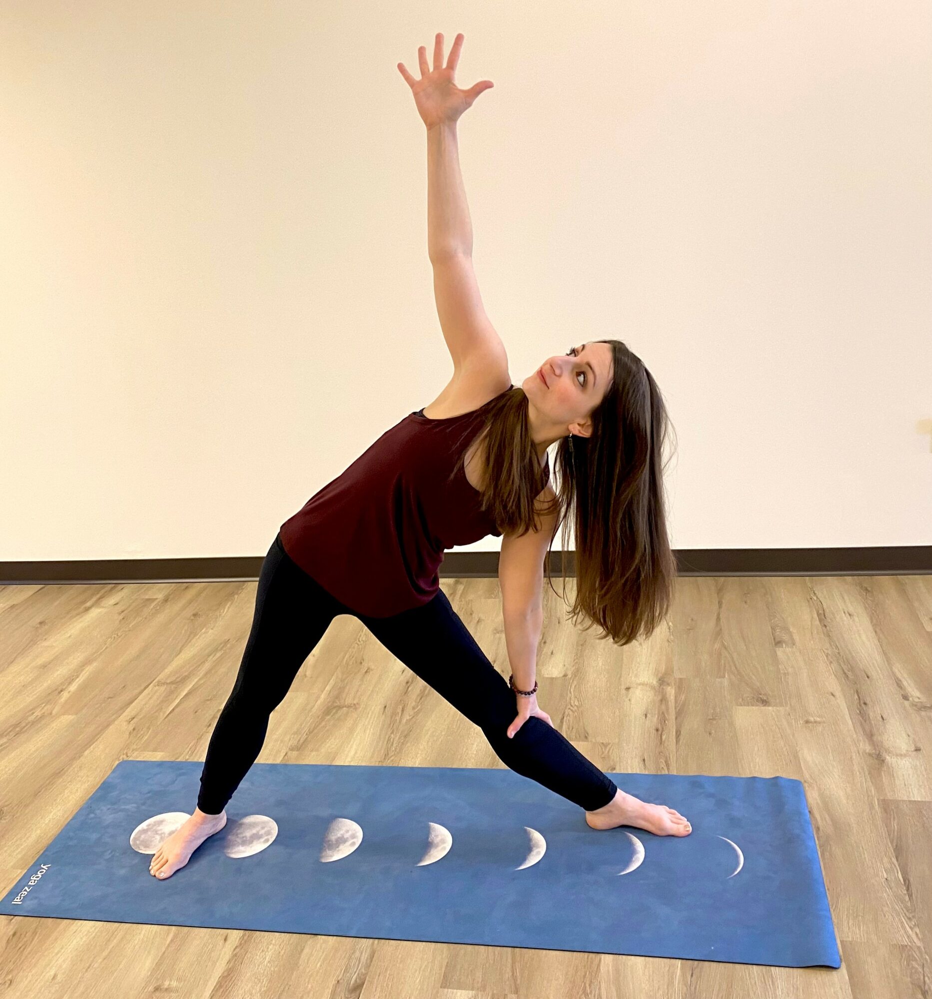 Woman practicing yoga on a blue mat, reaching upward with one arm, in a room with wooden floor and plain wall.