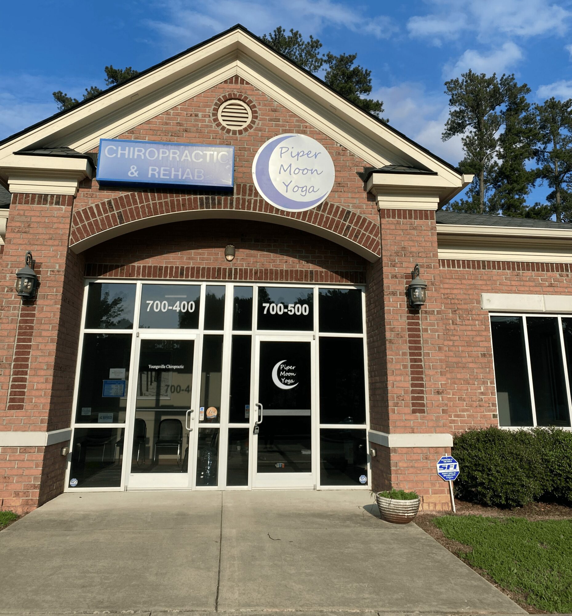 Brick building with glass doors, signs for chiropractic, Reiki, Moon Yoga, and therapy services, and a small bush outside.