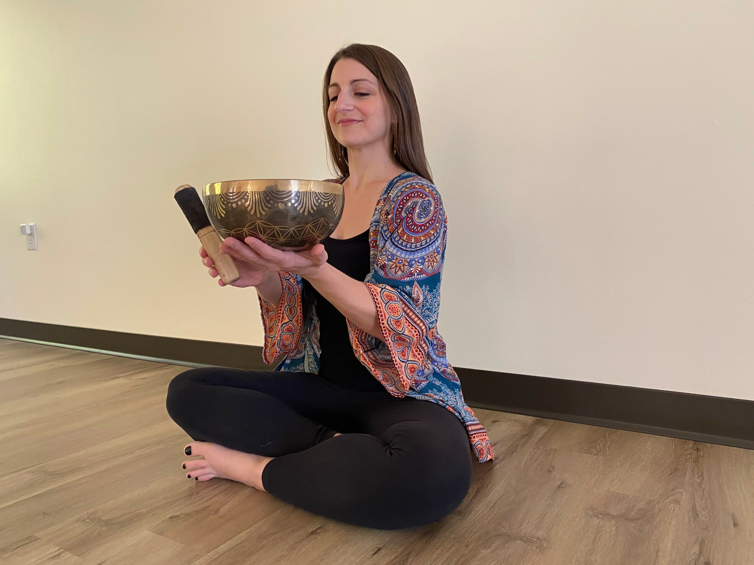 Woman sitting cross-legged on floor holding a large singing bowl, eyes closed, in a room with plain wall and wooden floor.
