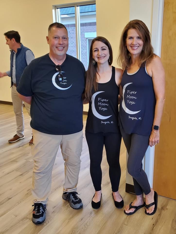 Three people standing indoors, smiling, wearing matching yoga tank tops, with a person in the background walking past.