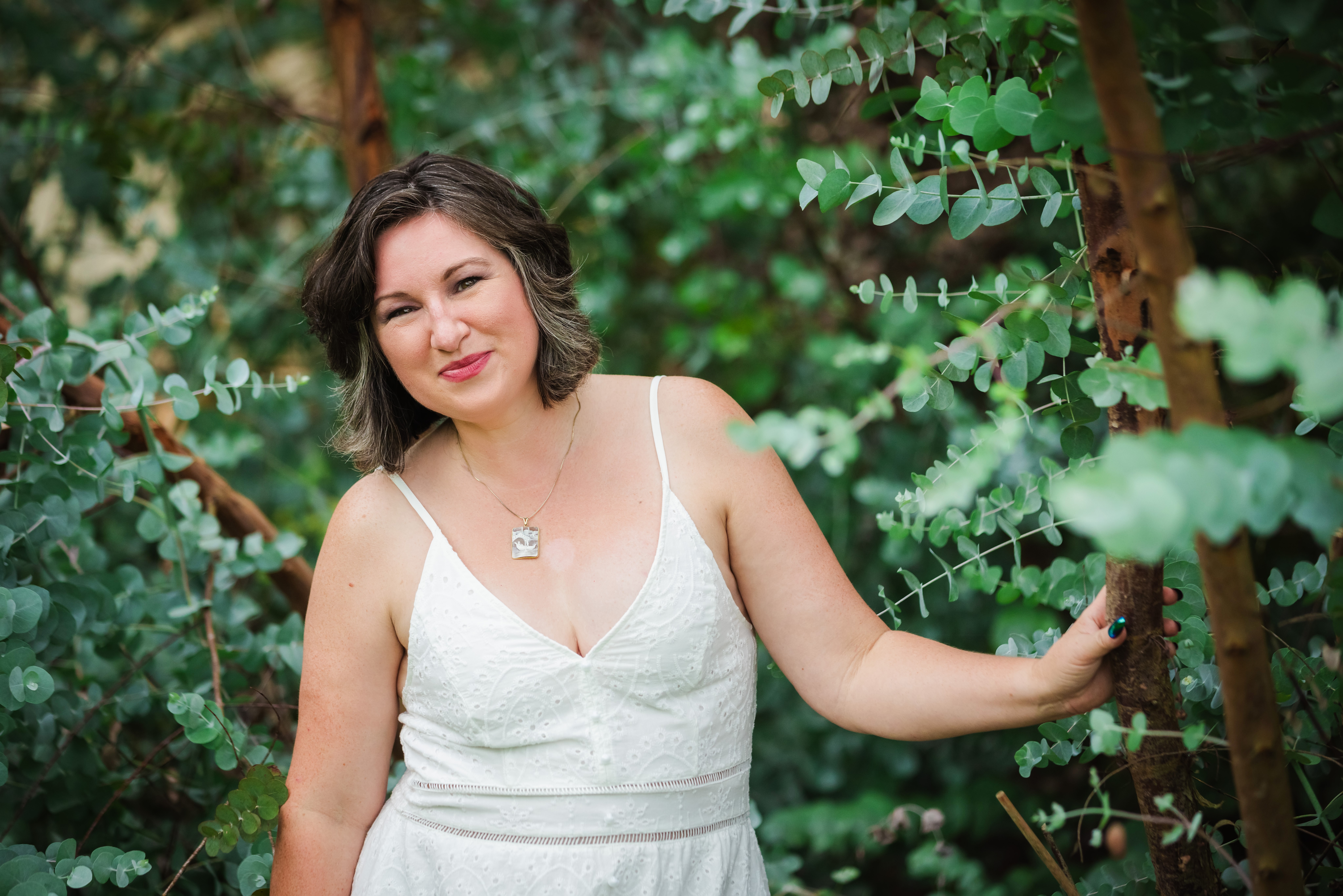 Woman with shoulder-length dark hair in a white dress standing outdoors among green trees and foliage.
