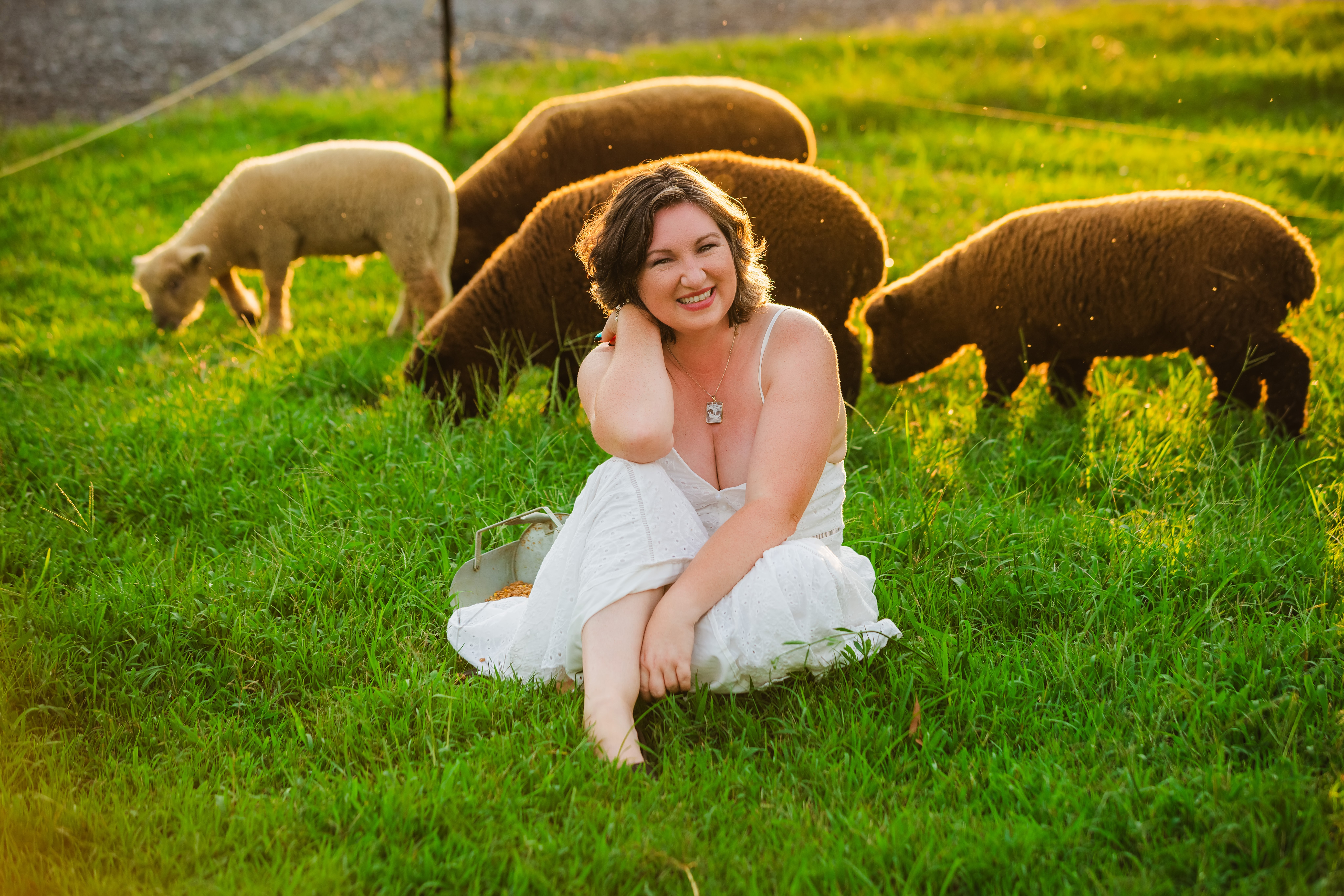 Woman sitting on grass with sheep grazing in the background, smiling and looking to the side.