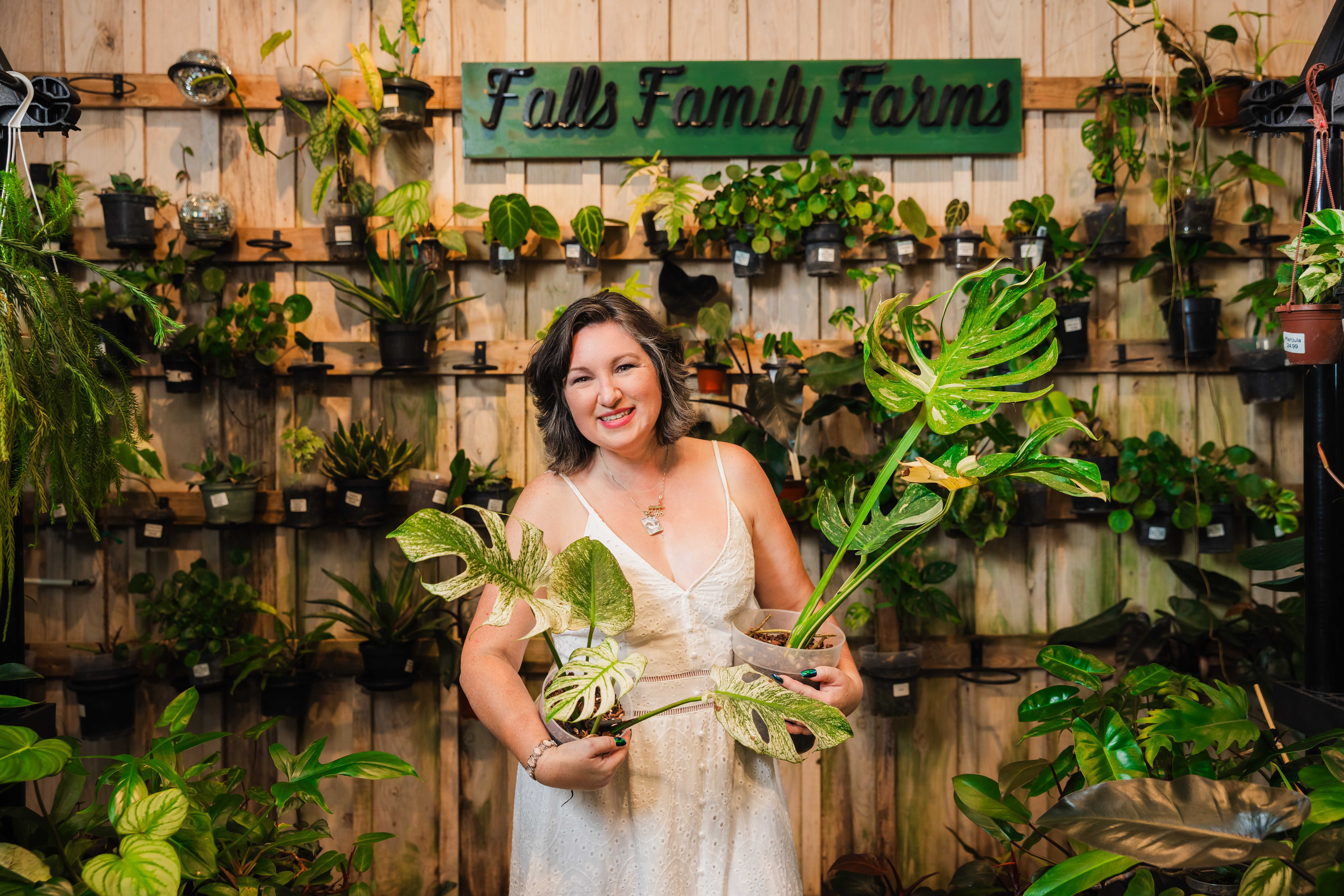 Smiling woman holding large green leaves in front of a wooden wall with potted plants and a sign that reads 'Fell Family Farm'.